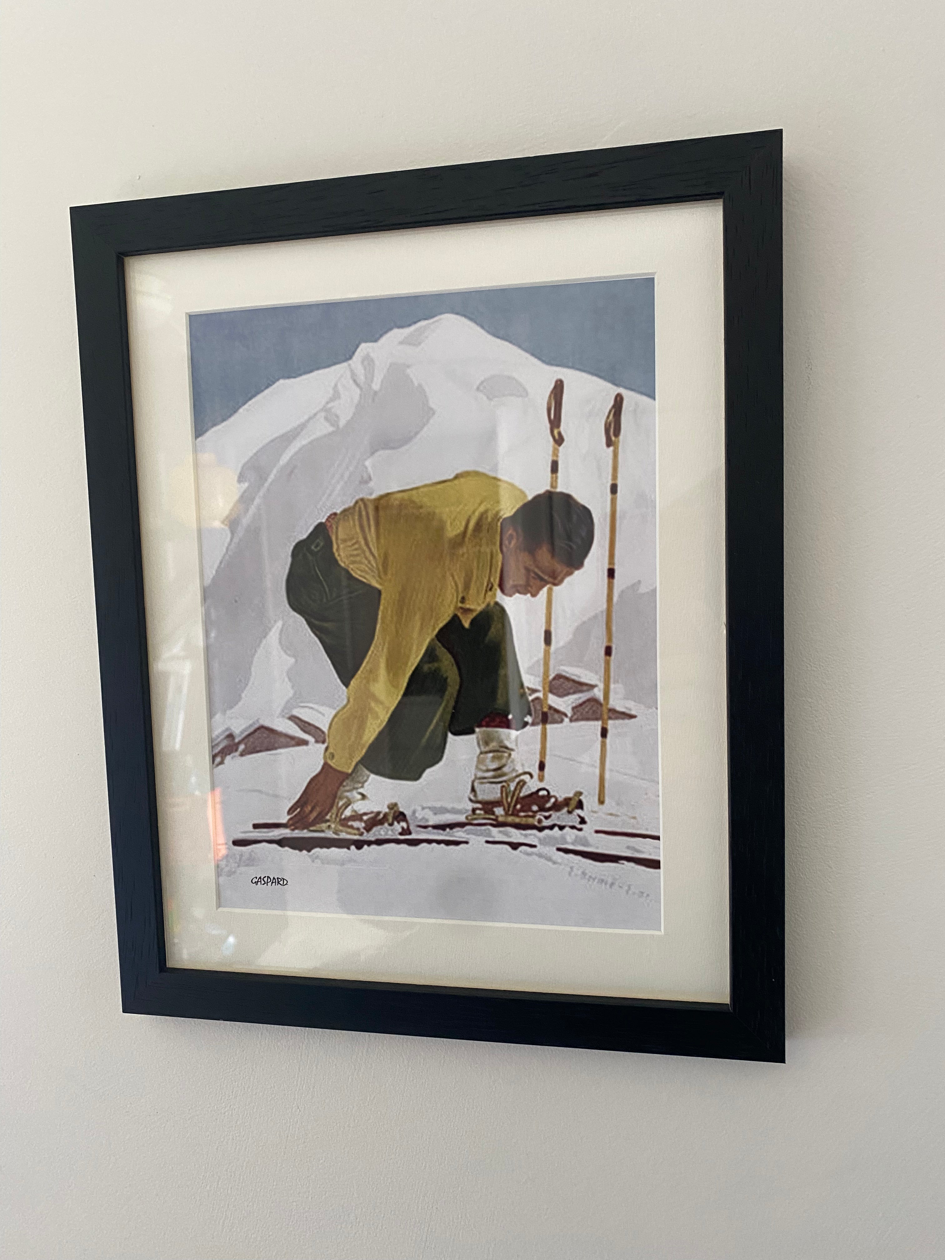 Black framed picture of a man in a yellow jumper & green ski pants leaning down to click into his wooden skis, with a white mountain & blue sky in the background, hanging on a white wall.
