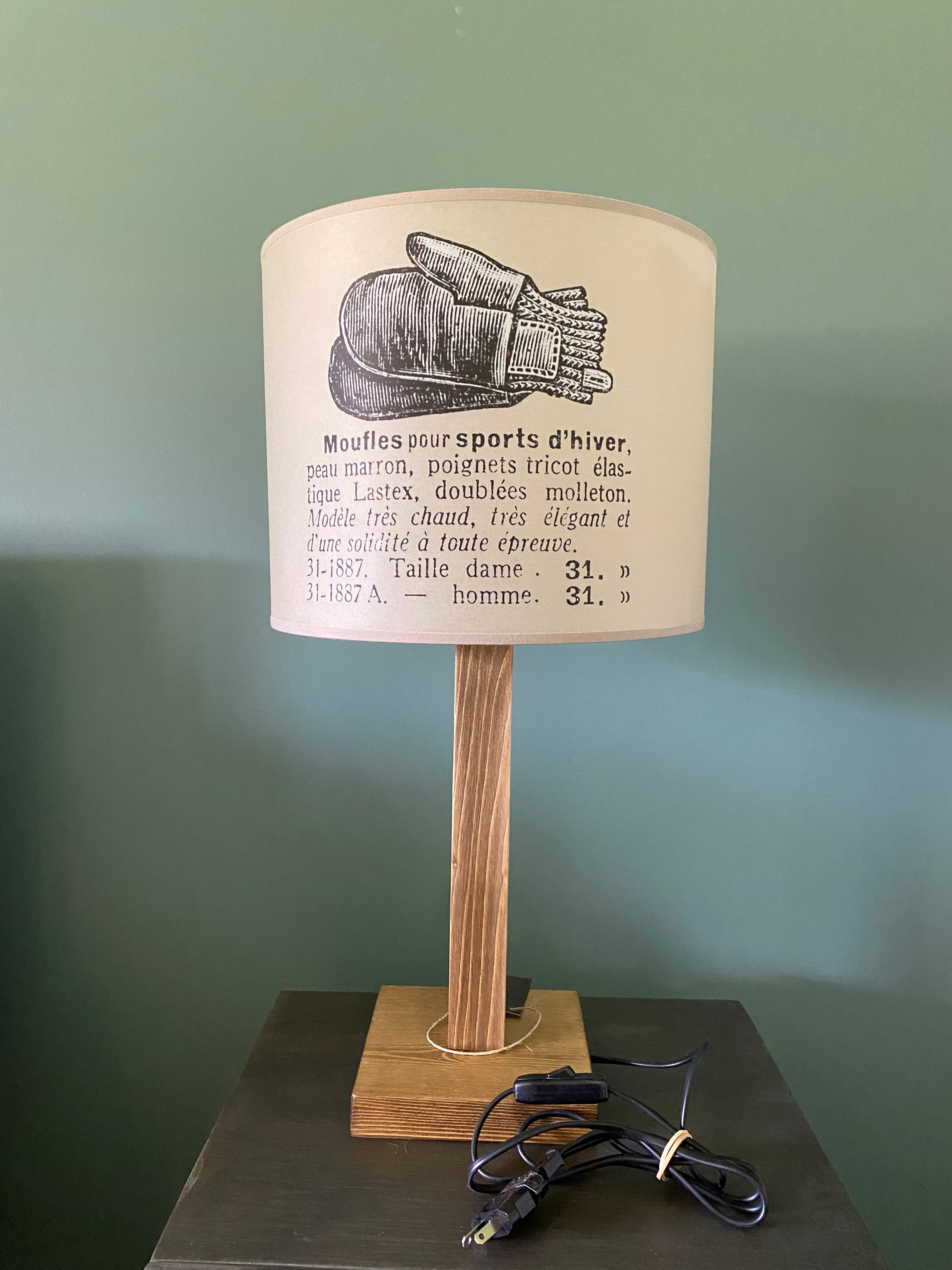Lamp with a square solid wooden base and square stem holding up the canvas printed beige lamp shade. The beige shade shows a vintage black & white drawing of a pair of woollen mittens and description in french of the mittens, showing the electrical cable. On a wooden table against a green wall.