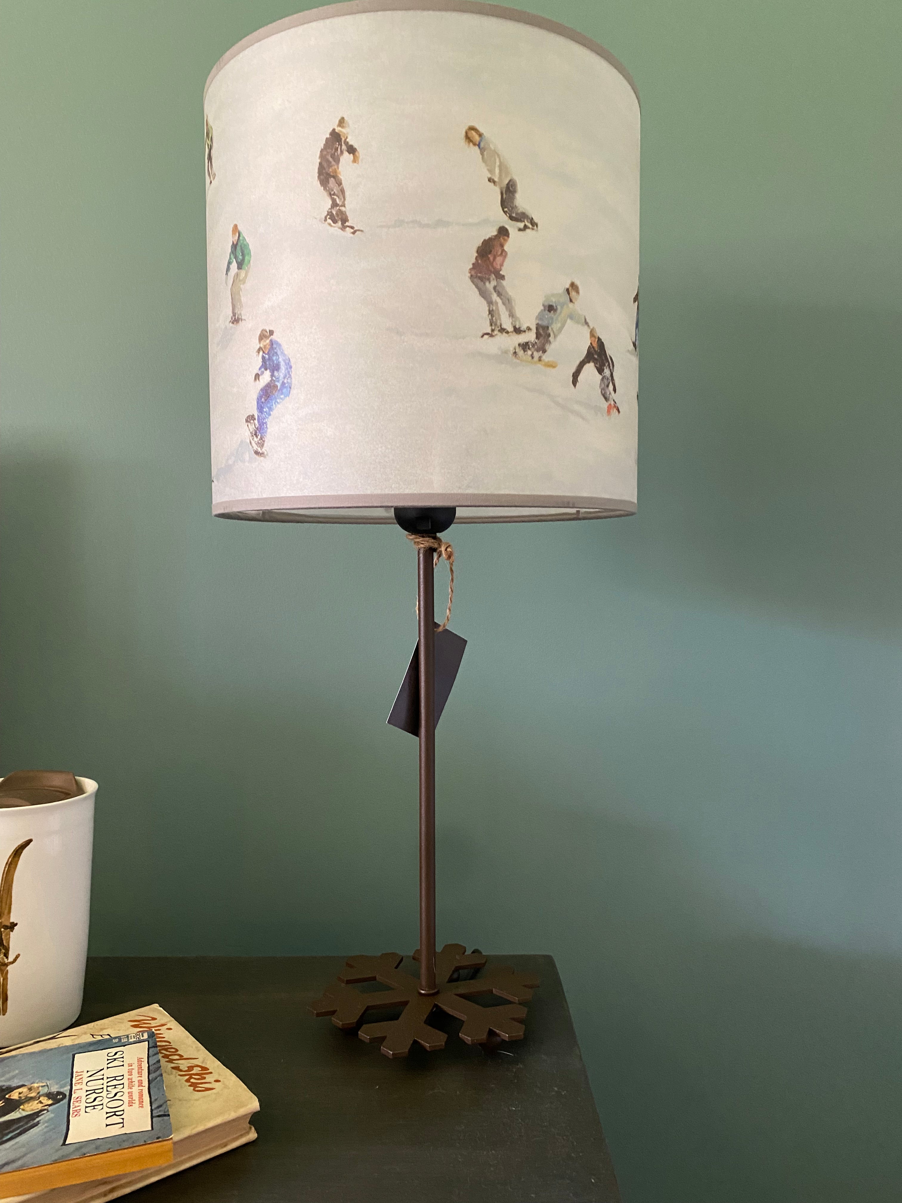 lamp with a round brown metal base in the shape of a snowflake, supporting a metal rod, topped by colour printed canvas lamp shade depicting a slope full of snowboarders riding downhill, in front a green wall & on top of a wooden table, with a coffee mug and two vintage books.