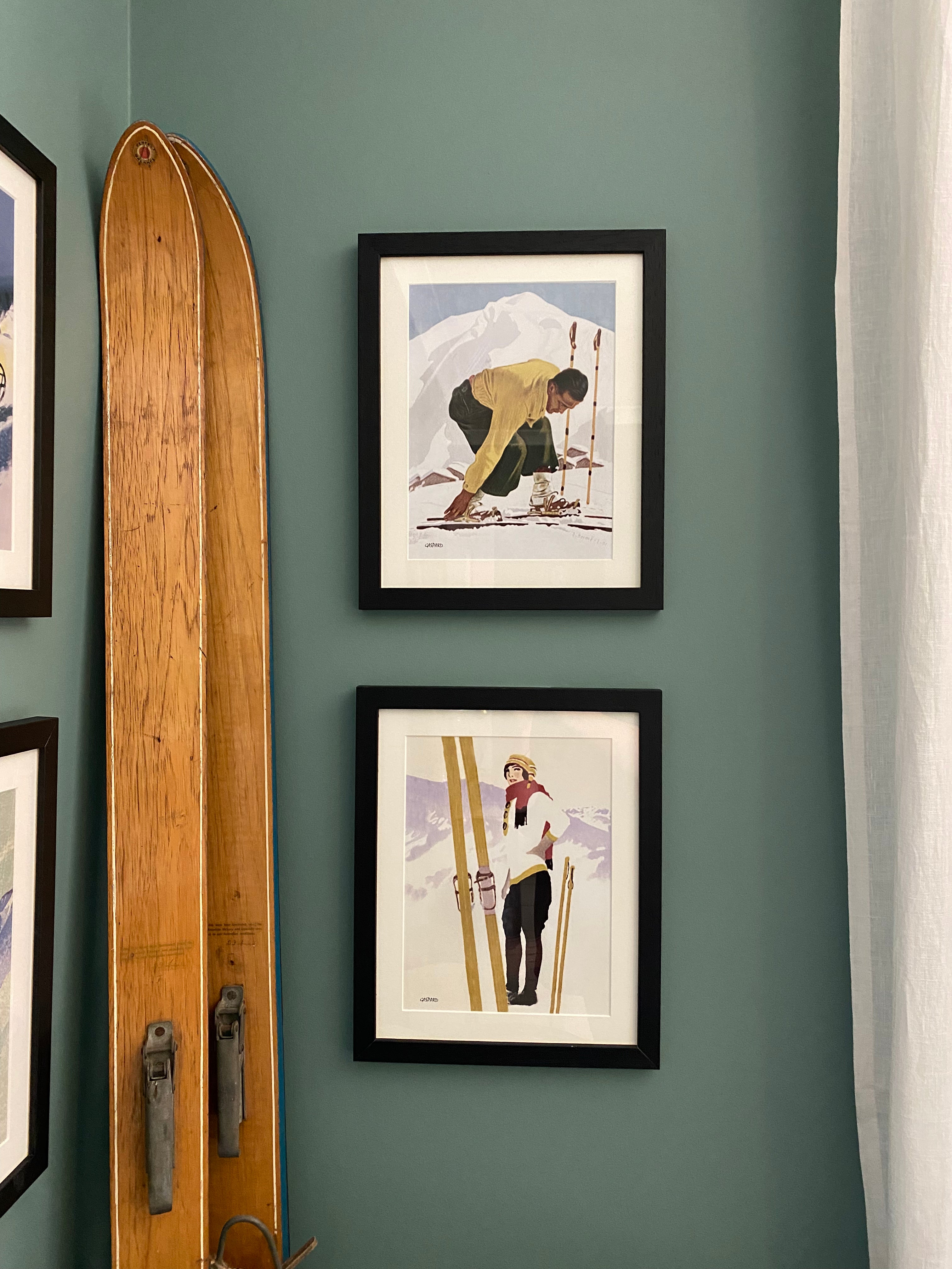 2 vintage black framed photos hanging on a green wall. 1: a woman standing in the snow next to a pair of wooden skis & poles upright in the snow in a white jumper, tan hat, tan gloves, a red scarf & black pants, with a pale sky casting a violet shadow across the mountains, 2; a man in a yellow jumper & green ski pants leaning down to click into his wooden skis, with a white mountain & blue sky in the background. Vintage wooden skis leaning against the wall 