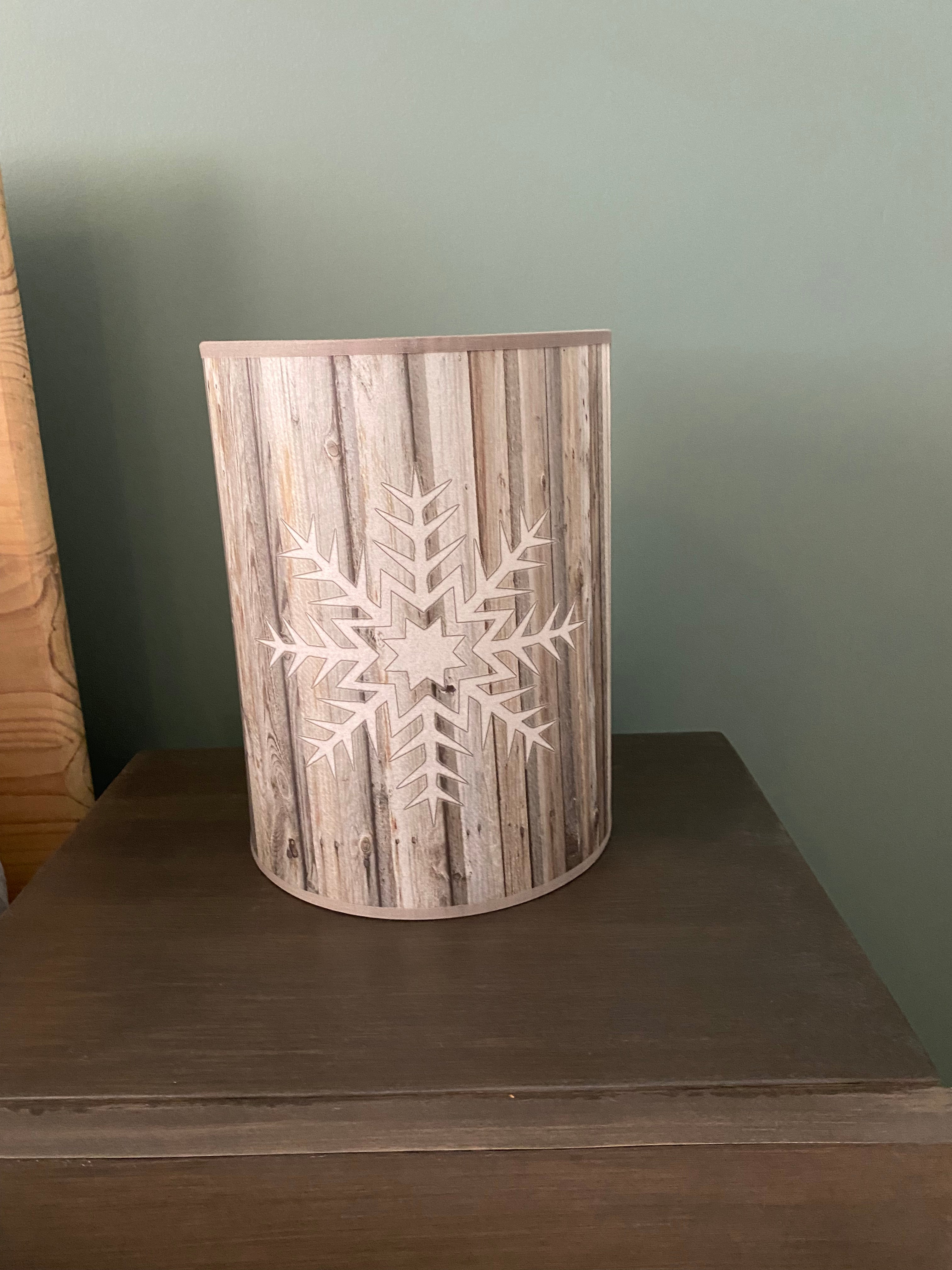 A  cylindrical table lamp made of a cylinder of canvas printed with a photo of wood grain, with a white snowflake embossed on opposing side of the lamp, against a green painted wall on a wooden table.