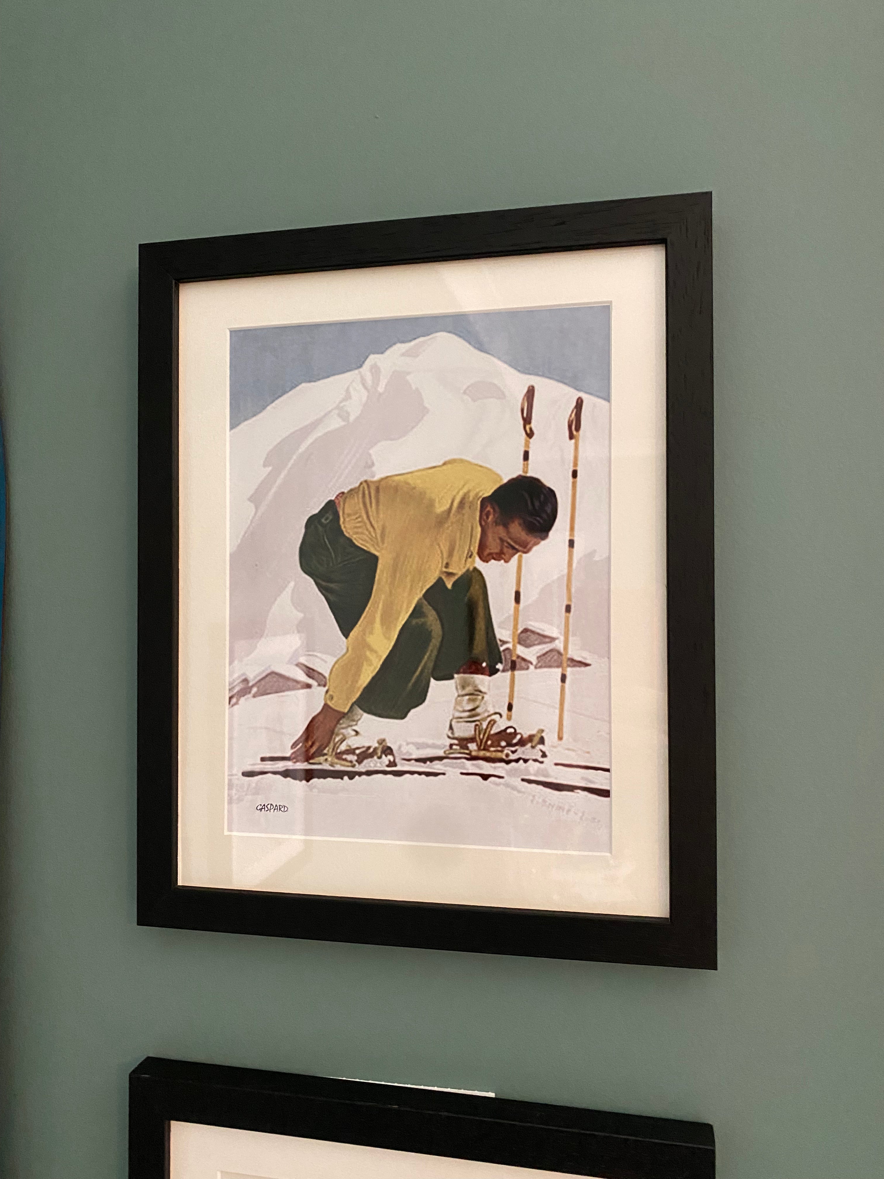 Black framed picture of a man in a yellow jumper & green ski pants leaning down to click into his wooden skis, with a white mountain & blue sky in the background, hanging on a green wall.