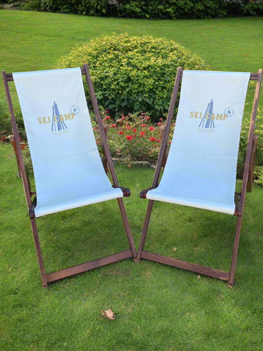 Front View of 2 Wooden deck chair frames with off-white canvas sling printed with an image of wooden skis stacked in a pyramid and the words ski camp written in yellow. 