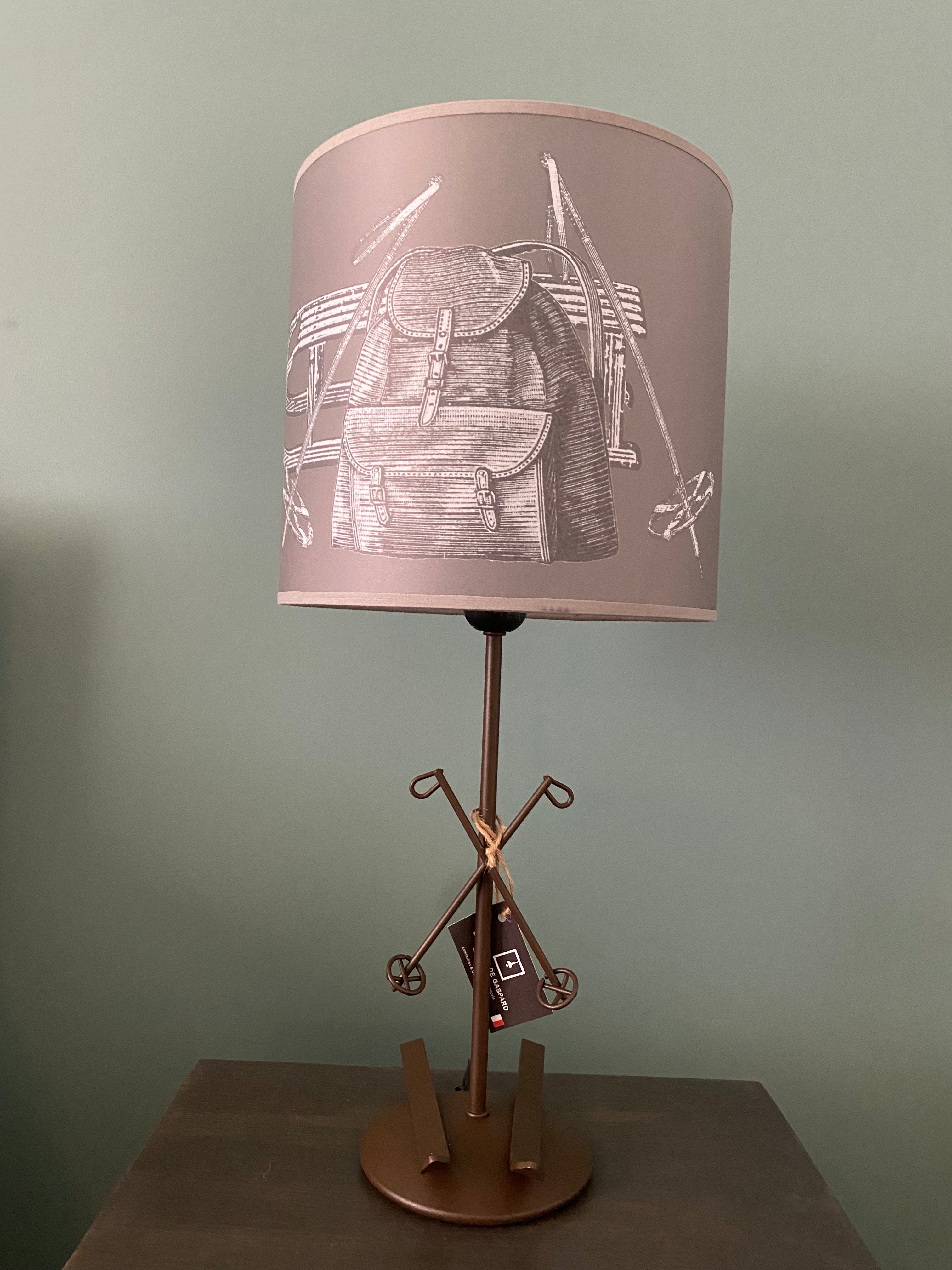 Lamp with a round brown metal base upon which rest a pair of skis supporting a brown metal rod with crossed metal ski poles mid way up the rod, topped by grey printed canvas lamp shade. The shade shows black and white drawing, against a grey background, of a pair of vintage snow goggles, a wooden sled, a pair of vintage ski poles and a vintage backpack. On a wooden table against a green wall.