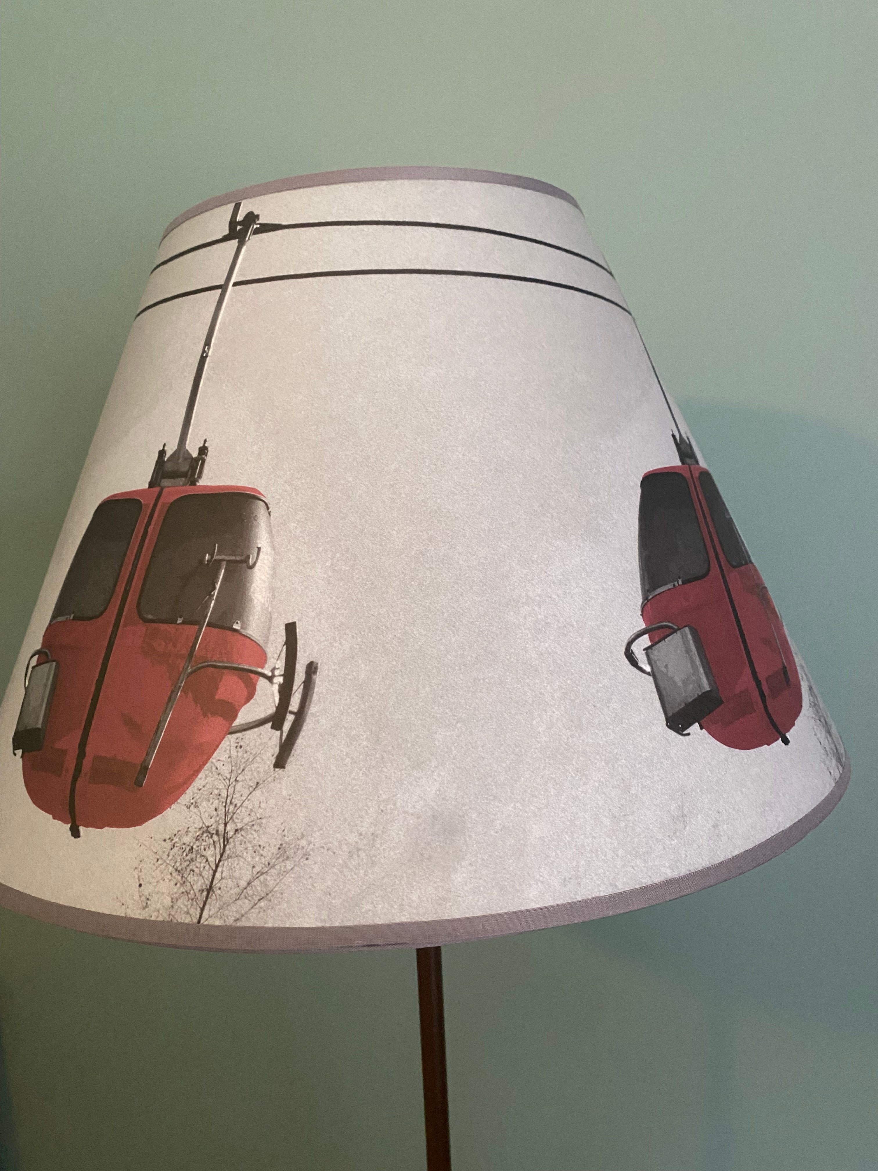Photo of a floor lamp with a brown metal rod, topped by a vintage photo printed canvas lamp shade. The colour photo shows 3 red egg gondolas in the air with tree tops and clouds in the background against a grey sky. Lamp in front of a green painted wall.