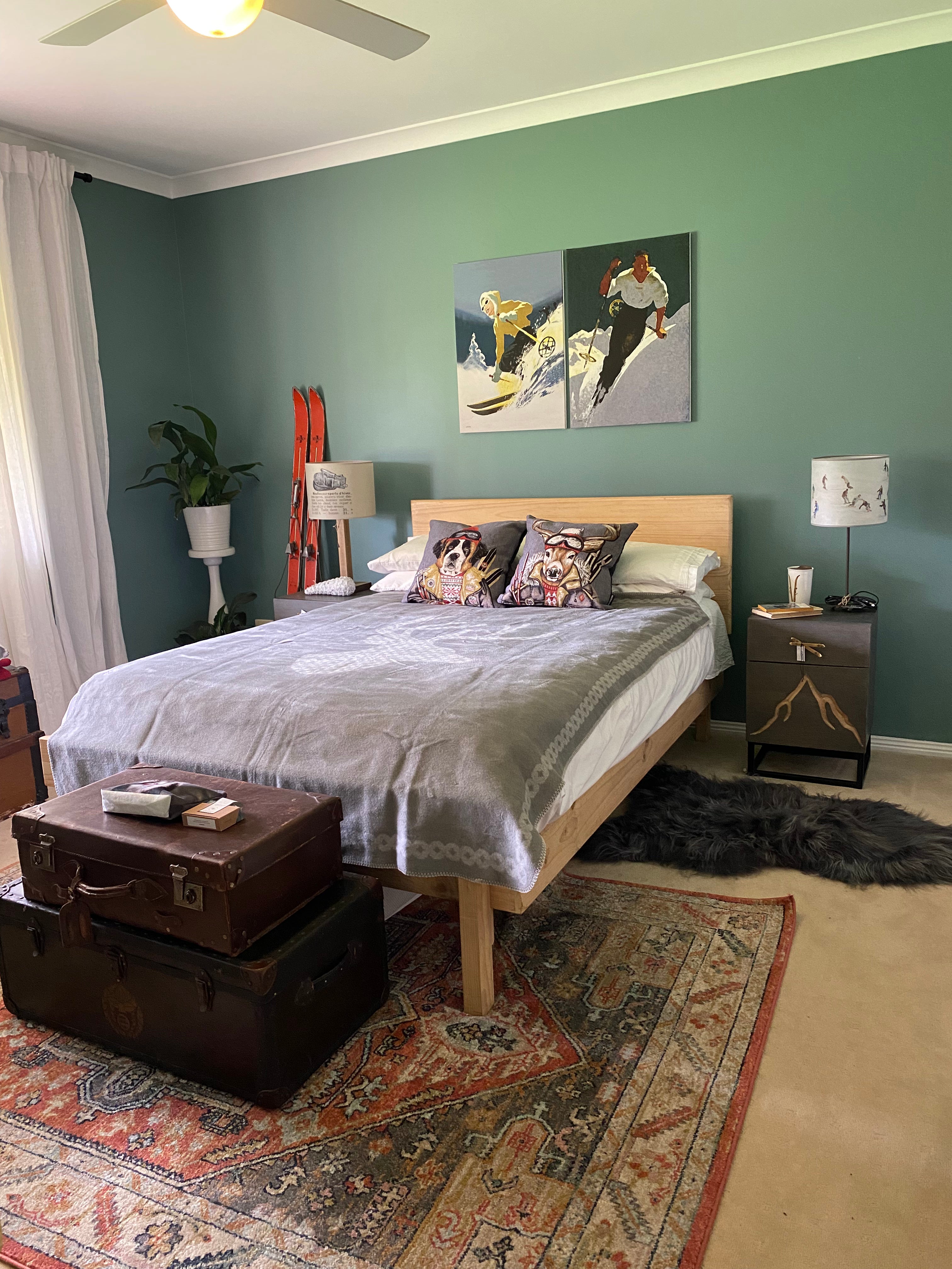 Wide angle view of a guest bedroom, with the Snowboarding lamp on the right hand bedside table. Room painted green, with vintage suitcases at the end of the bed and vintage orange skis leaning against the wall. Grey Deer blanket on the bed and two throw cushions