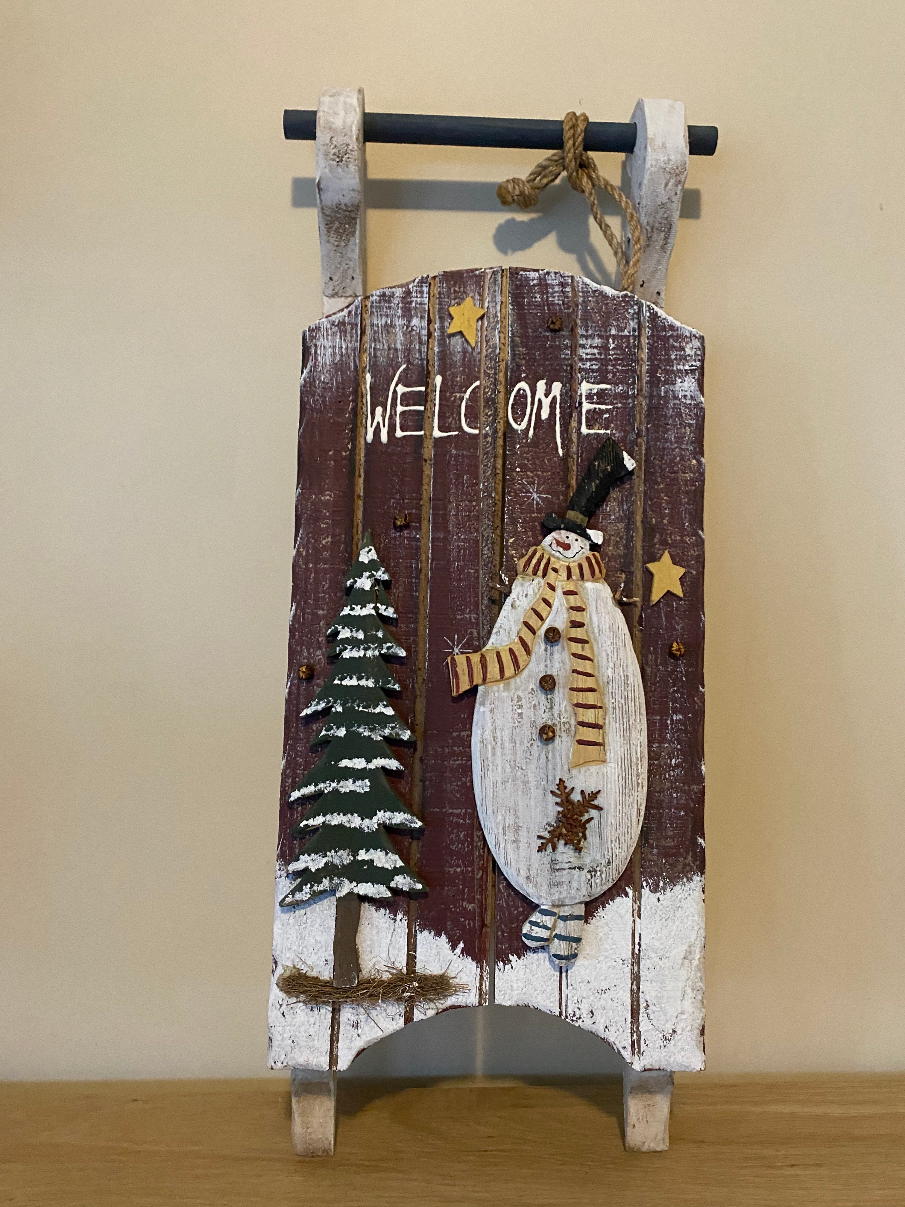 wooden decorative welcome sign in the shape of a vintage sled, decorated with a snowman and a snow-covered pine tree. Resting against a beige wall, on a wooden table. front view