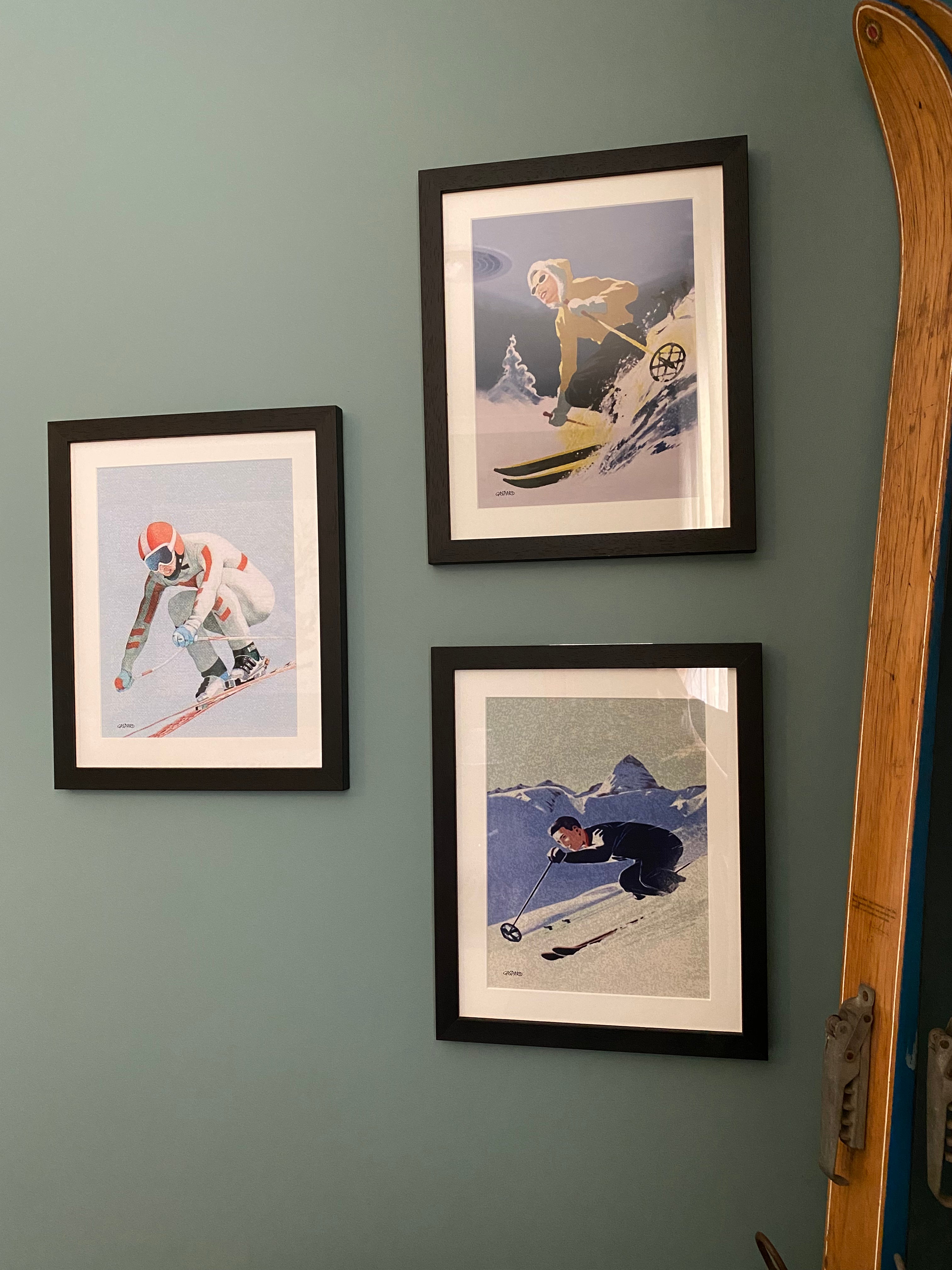 3 vintage black framed photos hanging on a green wall. 1st is Black framed vintage photo of a woman in a yellow jacket with a white fur at the edge of the hood and blue pants skiing downhill, leaning to the right into the hill with a tree and the shadow of the mountains in the background below a blue sky. Second is a man in blue suit skiing aggressively downhill, with mountains behind him. Third is a crouching ski racer, light blue sky behind. Vintage wooden skis leaning against the wall on the right