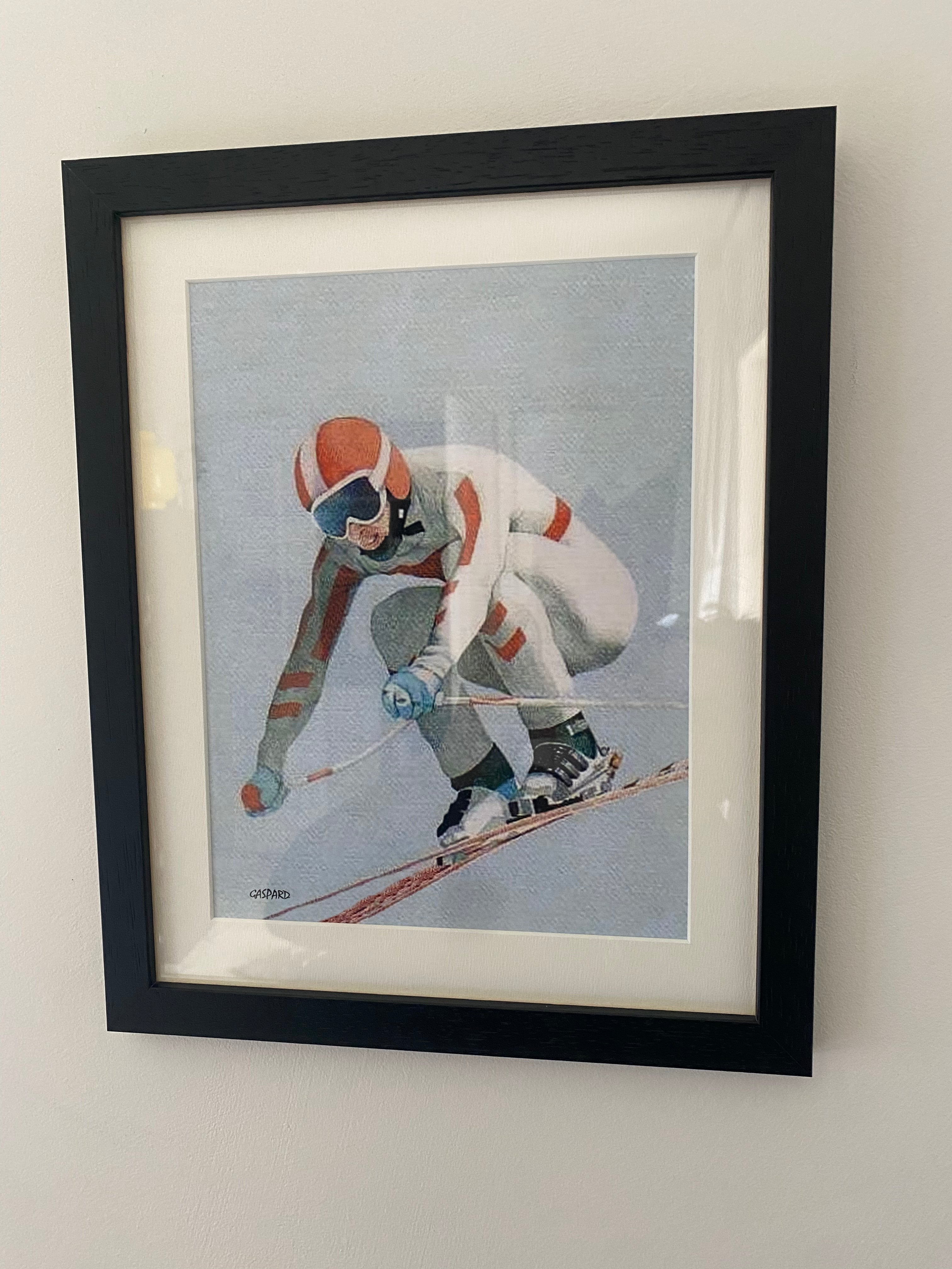 Black framed print of a person in a white fitted racing suit with red stripes, a red helmet with a central white stripe, with skis, boots and poles in a crouched position airborne and skiing downhill, with a light blue sky in the background, hanging on a white wall.