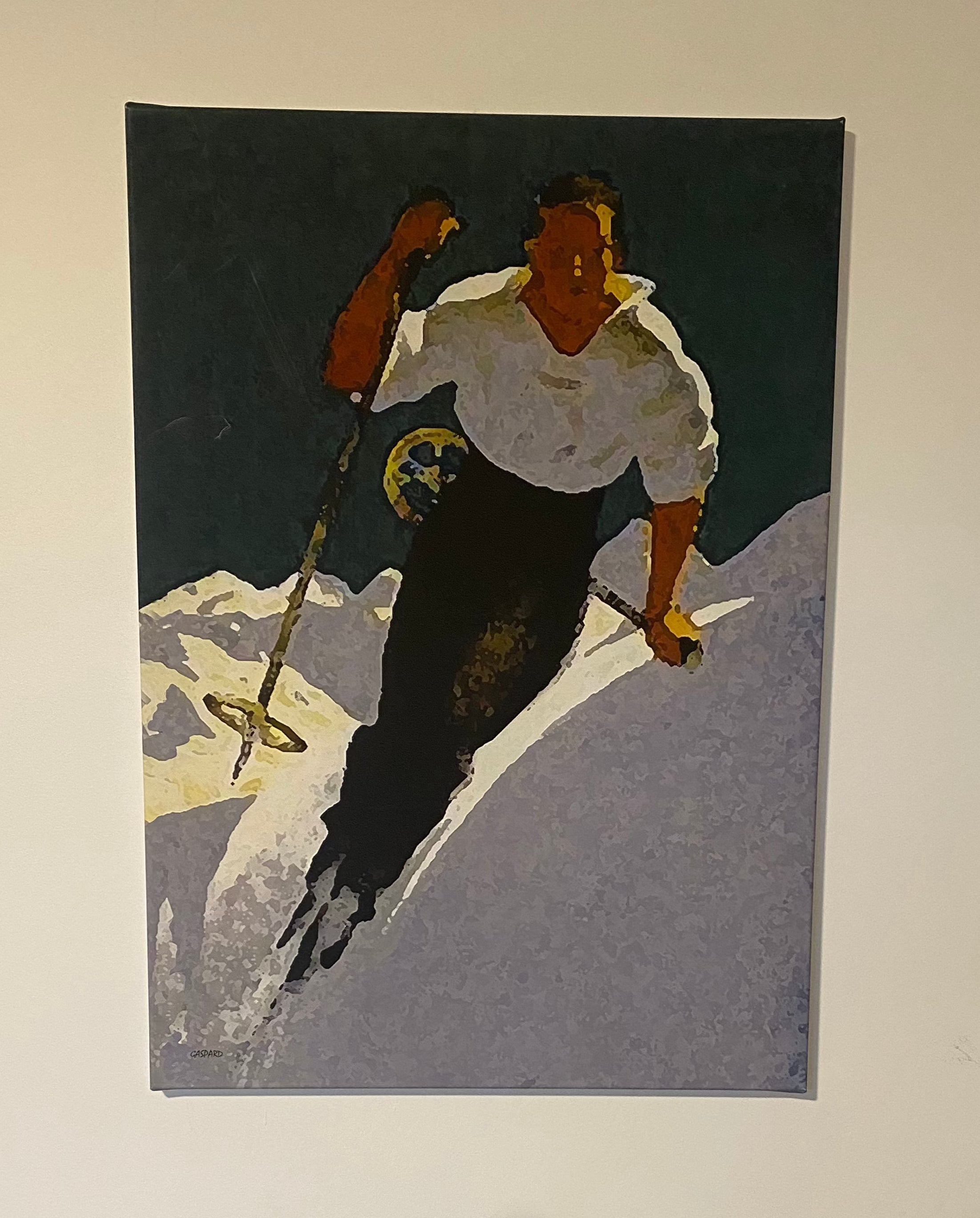 Canvas print of a man in a white shirt and blue pants skiing downhill, leaning left into the hill with mountains in the background below a dark green-blue tinged sky.  Hanging on a white wall