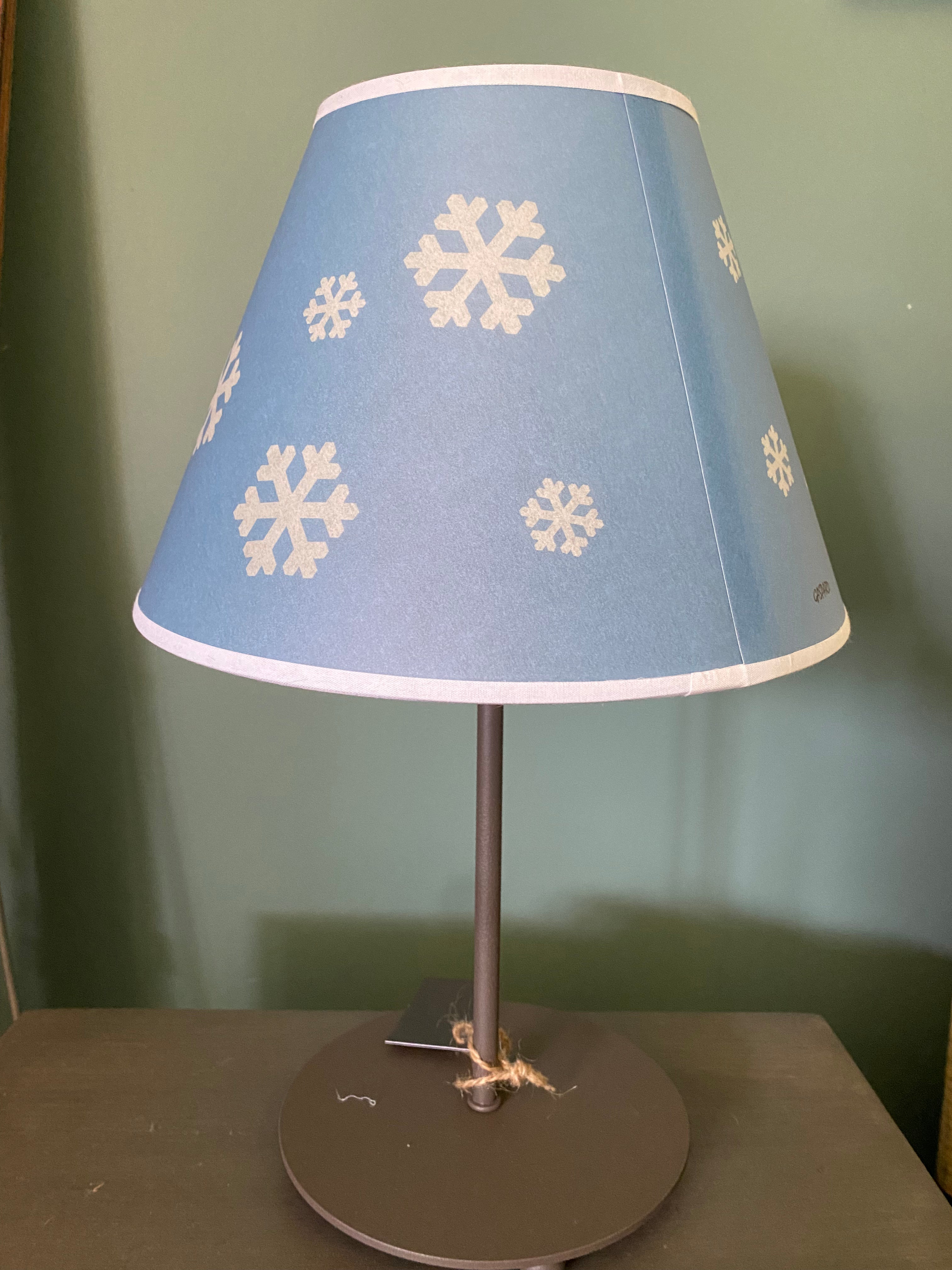 lamp with a round brown metal base supporting a metal rod, topped by snowflake printed canvas lamp shade in blue and white. Sitting on a wooden table against a green painted wall.