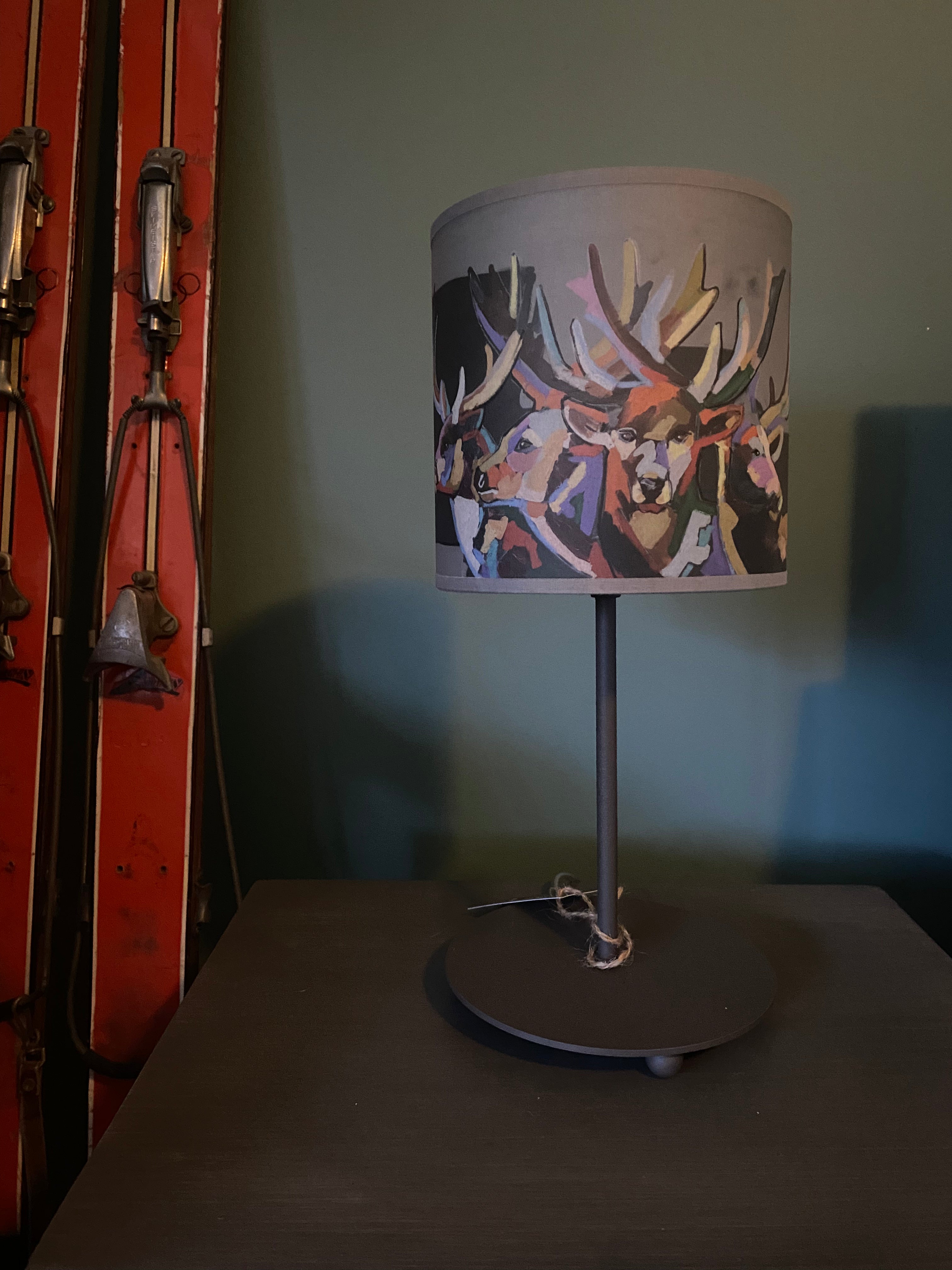 Lamp with a round brown metal base, with simple rod supporting the light shade, that shows multiple deers rendered gorgeously in multiple colours, against a green wall, sitting on a wooden table with a pair of orange vintage skis on the left hand side of the table.