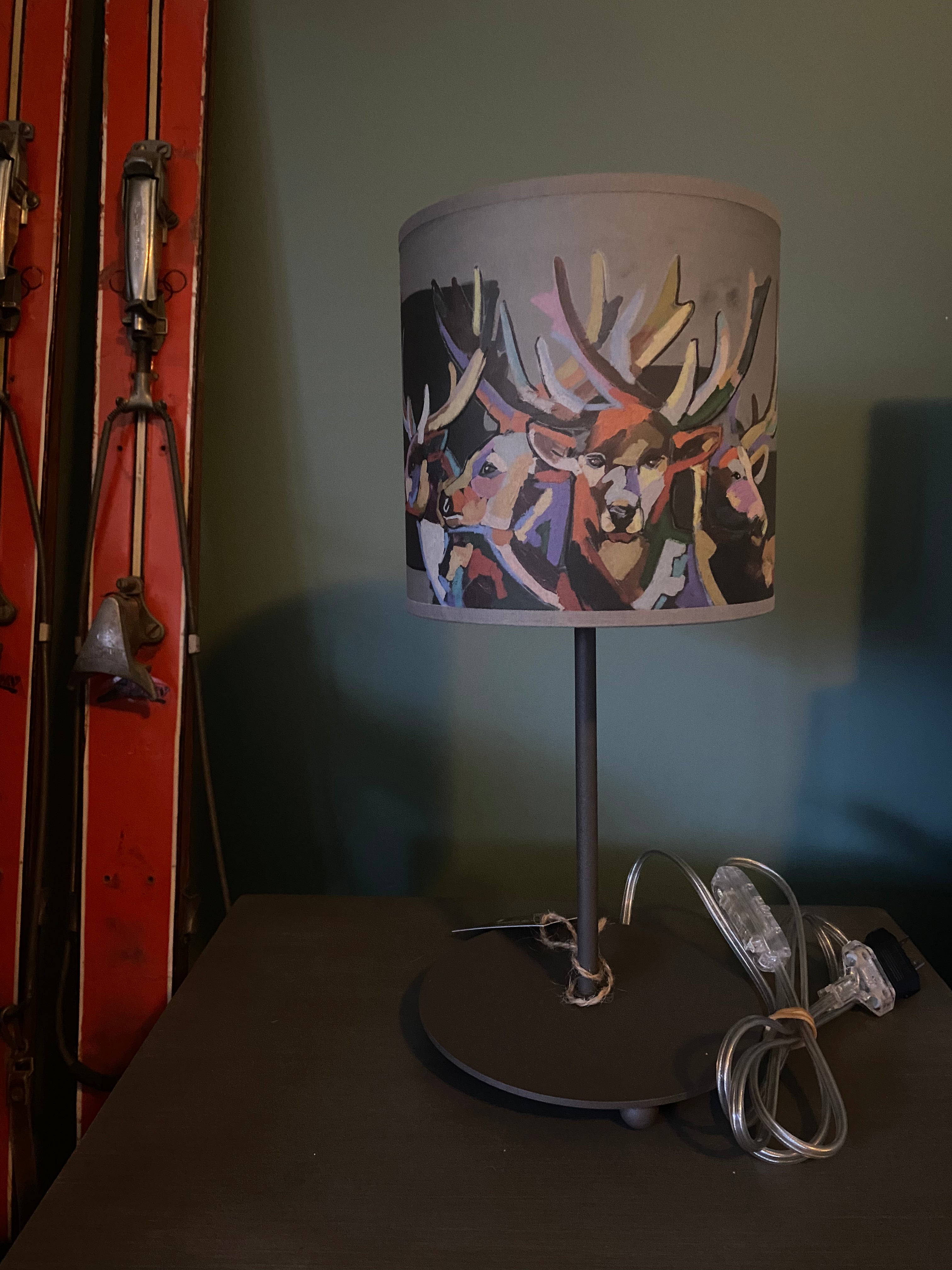 Lamp with a round brown metal base, with simple rod supporting the light shade, that shows multiple deers rendered gorgeously in multiple colours, with cord shown. In the background is a green wall, sitting on a wooden table with a pair of orange vintage skis on the  left hand side of the table.