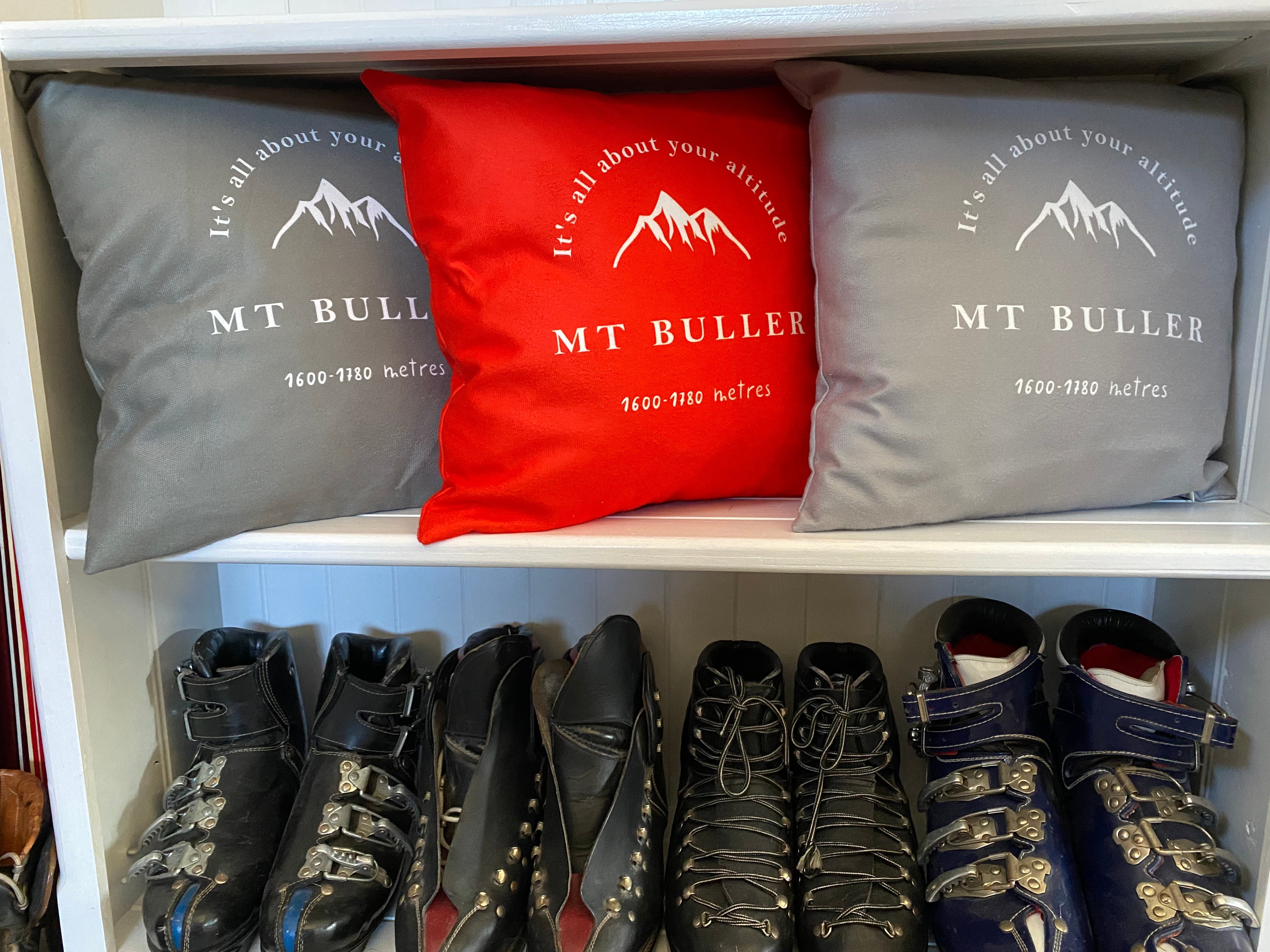 Image of white bookshelf with red, grey and taupe cushions printed with the words Mt Buller; it's all about your altitude visible on the cushion and a drawing of a mountain, and vintage leather ski boots underneath