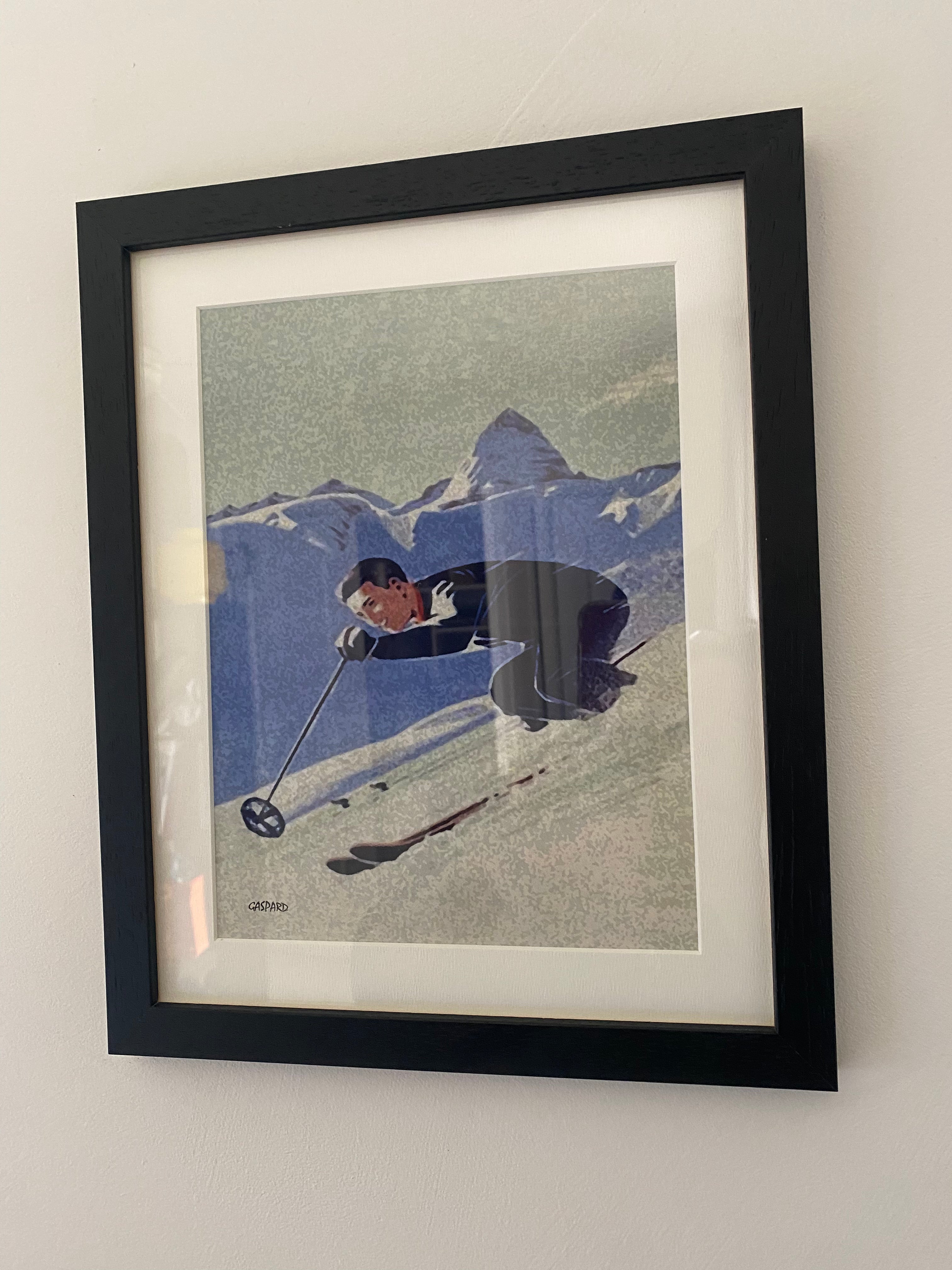Black framed picture of a man in blue suit skiing downhill, with shaded mountains in the background below a green-blue tinged sky, hanging on a white wall.