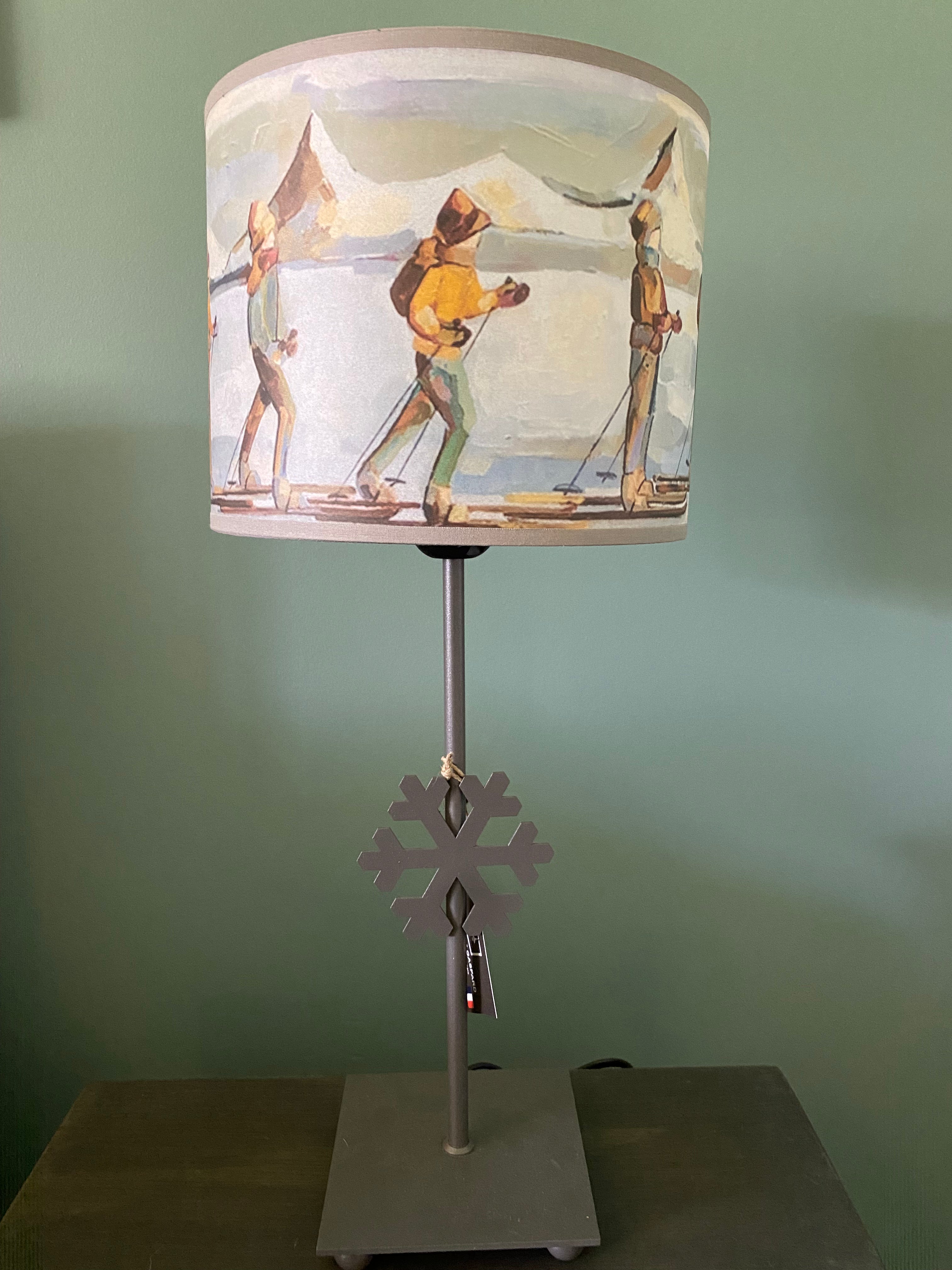 lamp with a square grey solid metal base supporting a rod with ca snowflake mid way up the rod, topped by a colourful canvas lamp shade. The shade depicts people on skis skinning across the snow in a line, one in front of the other, with snow capped mountains in the background. Sitting on a wooden table behind a green wall. Different view of shade, again