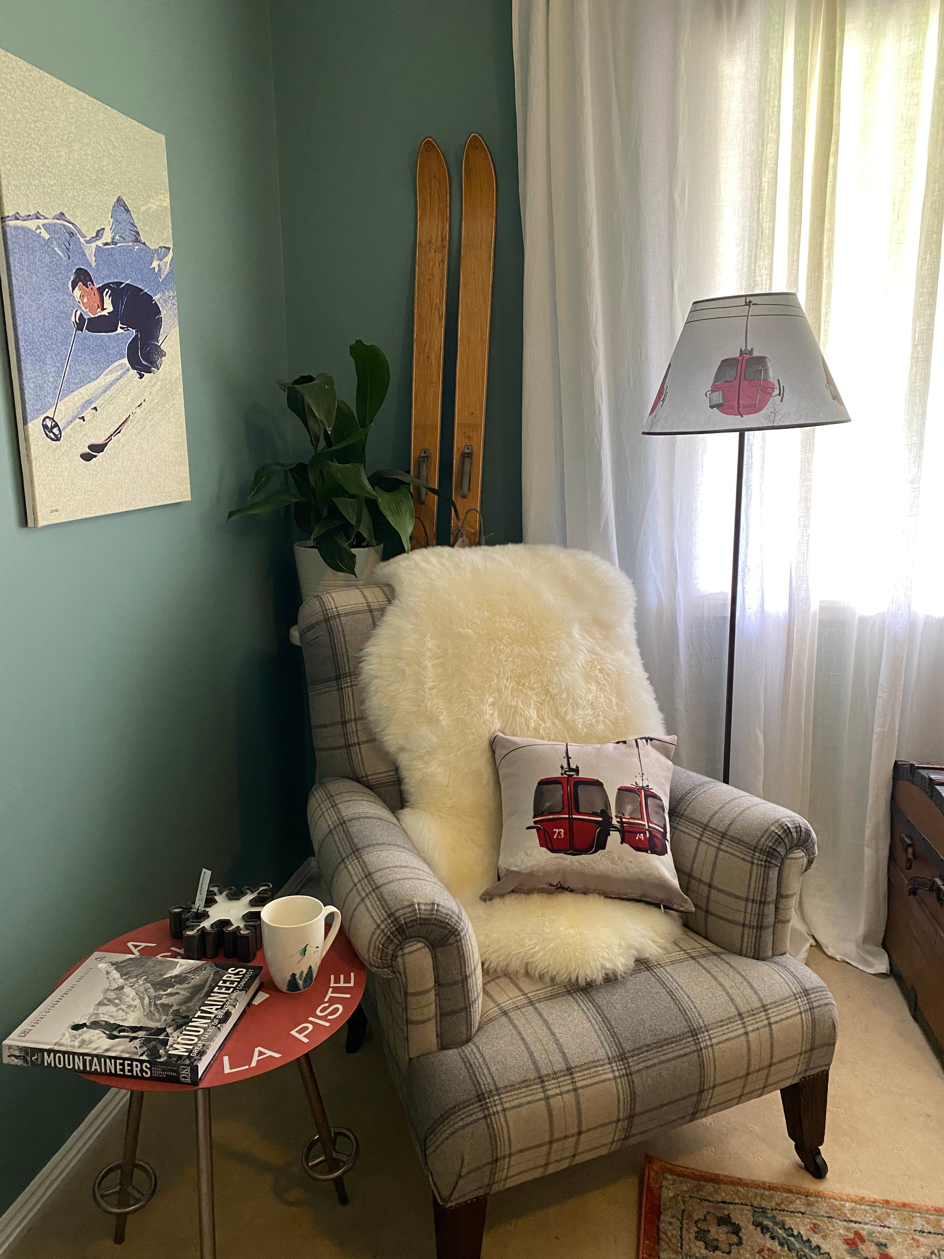 Photo of a blue tartan armchair, white sheepskin rug & red gondola cushion, with lamp & vintage trunk to the right, pot plant & wooden skis behind the chair, vintage skiing print on the left wall & alpine resort sign table with a book, a mug and candle on the left of the chair. Floor lamp with a square solid wood base supporting a brown metal rod & conical a lamp shade. Shade: 3 red egg gondolas in the air with tree tops and clouds in the background. Beige carpet, a green painted wall, white curtains.