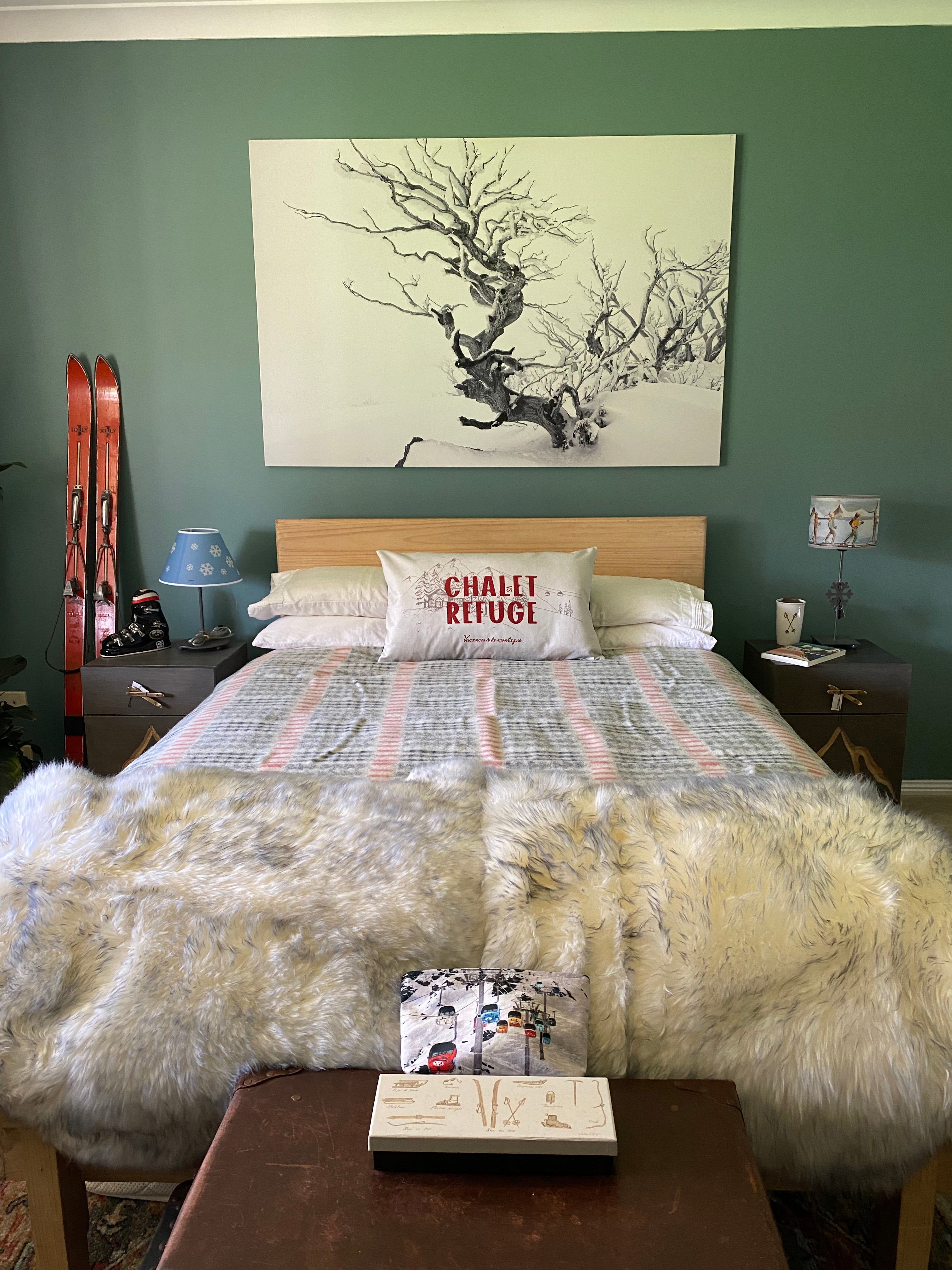 Front on view of a bedroom, with the Snowflake lamp on the right hand bedside table. Room painted green, with vintage suitcases at the end of the bed and vintage orange skis leaning against the wall. Grey, red & white blanket on bed & 1 throw cushion