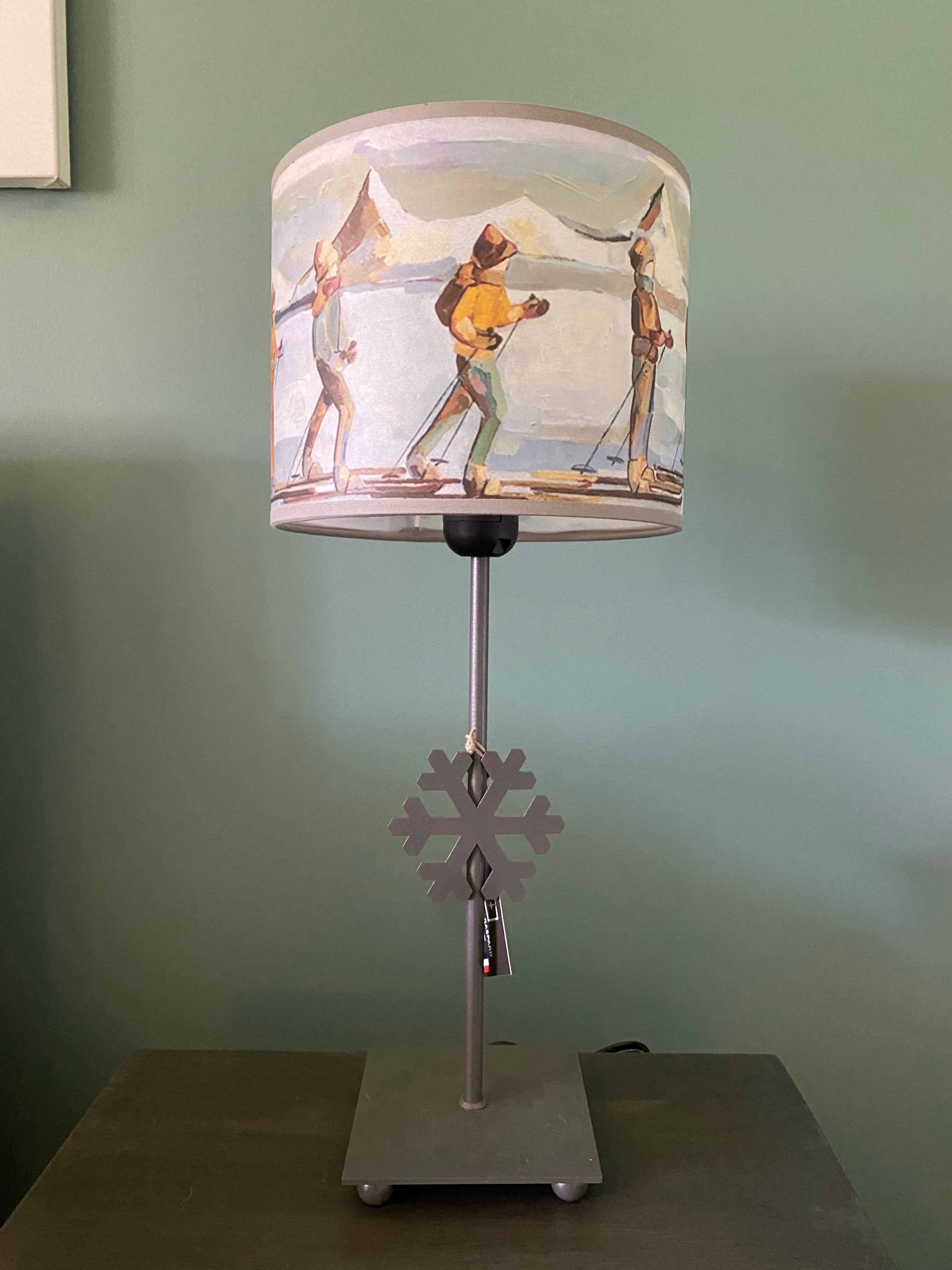 lamp with a square grey solid metal base supporting a rod with ca snowflake mid way up the rod, topped by a colourful canvas lamp shade. The shade depicts people on skis skinning across the snow in a line, one in front of the other, with snow capped mountains in the background. Sitting on a wooden table behind a green wall. Different view of shade.