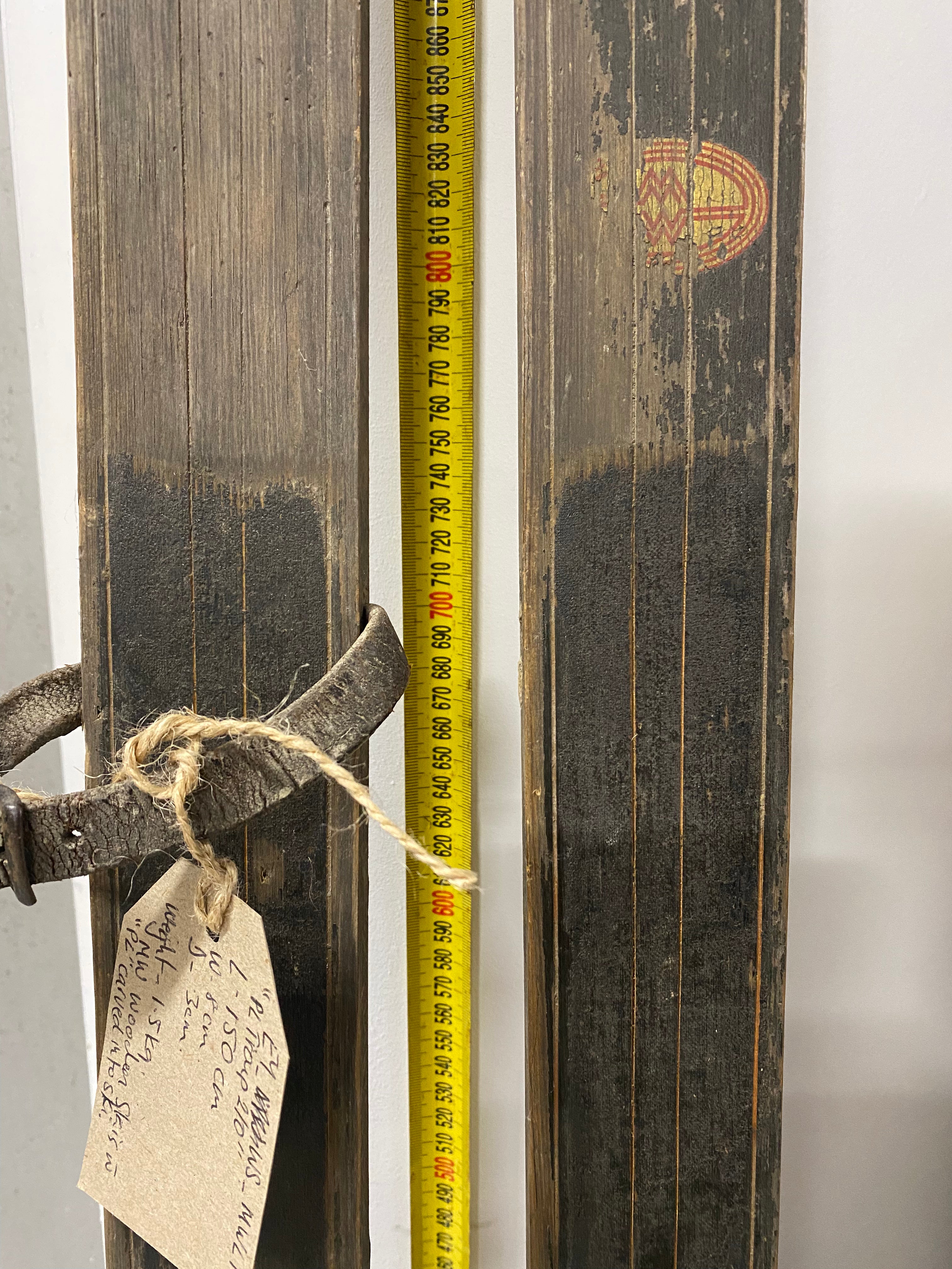 Front view of ski bindings: Montgomery Ward vintage wooden skis. 1 skis with a leather strap. 1 ski sun faded. leaning against white painted wall with yellow measuring tape