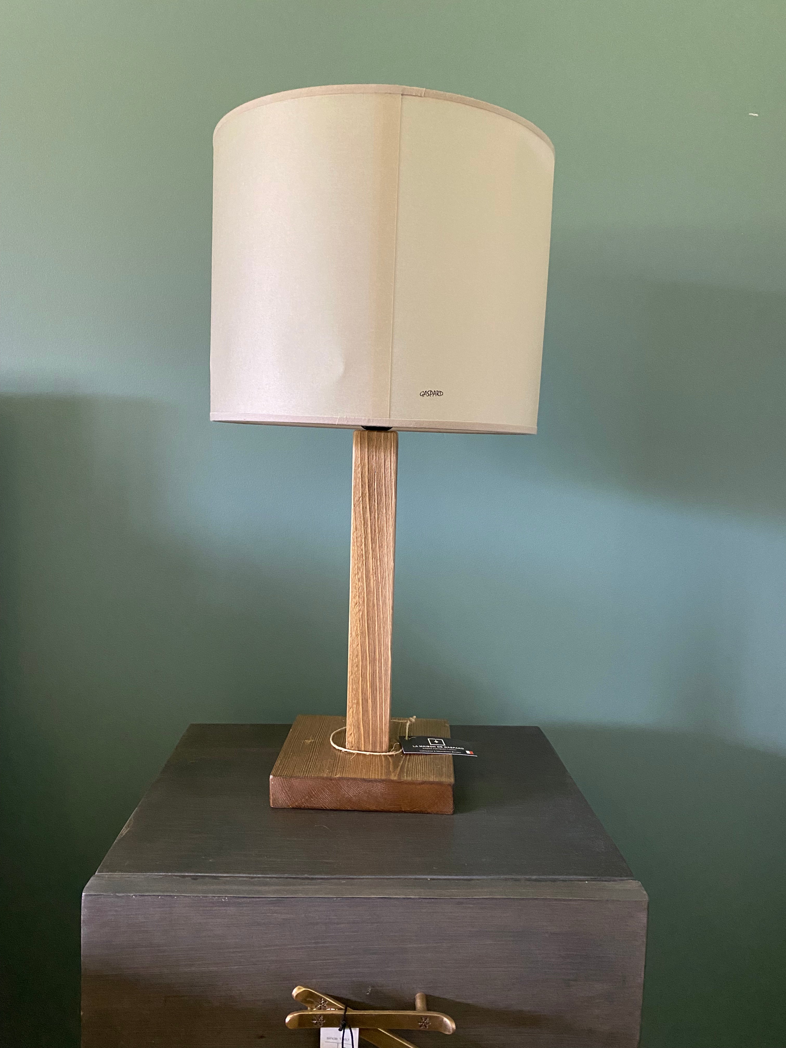 View of the back of the lamp and shade. Lamp with a square solid wooden base and square stem holding up the canvas printed beige lamp shade. The back of beige shade is blank. On a wooden table with brass ski handles against a green wall.