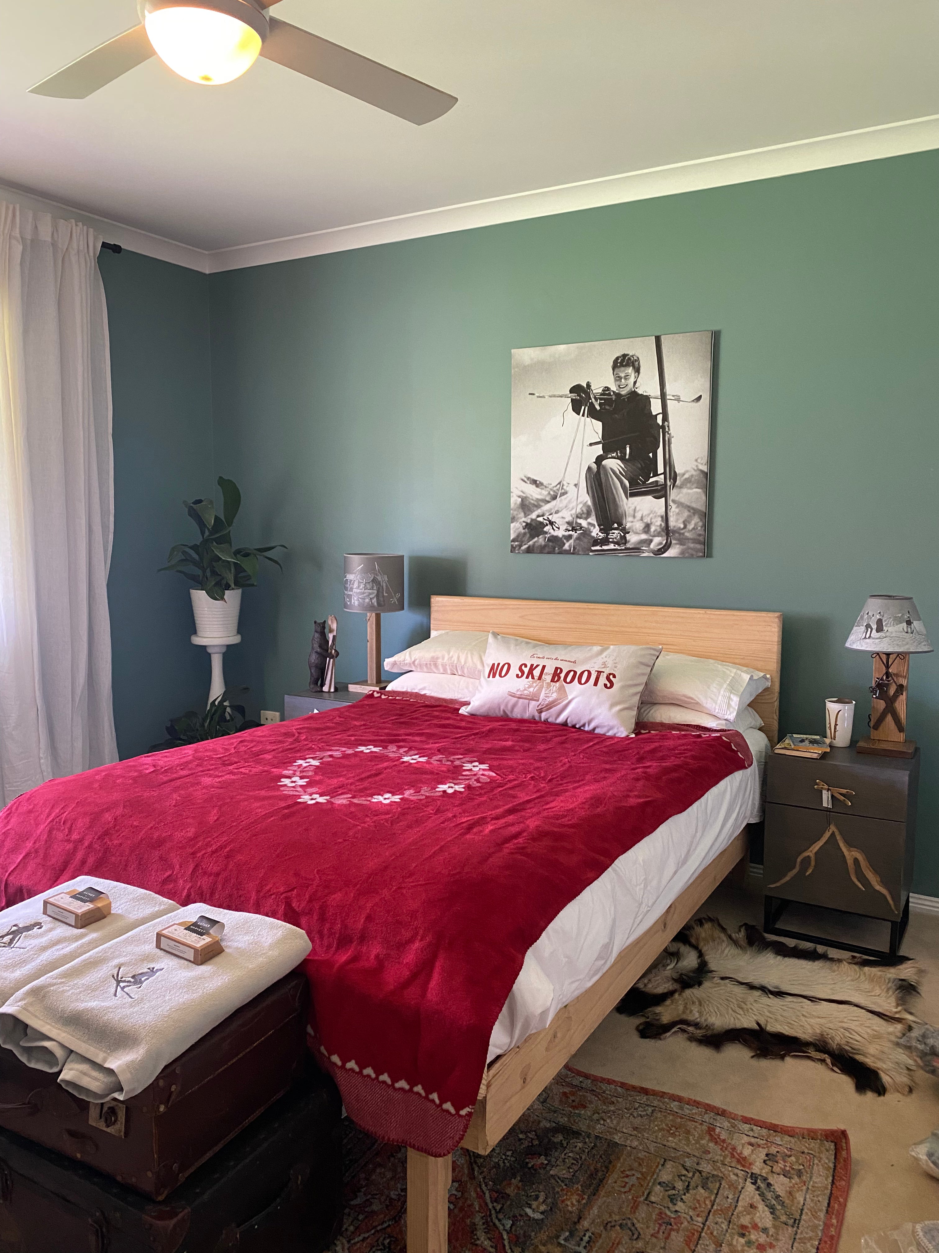 Wide angle view of a guest bedroom, with the Wooden Vintage Ski & Pole lamp on the right hand bedside table. Room painted green, with two towels and soaps resting on vintage suitcases at the end of the bed, a pot plant in the far corner, red blanket on the bed and one throw cushion