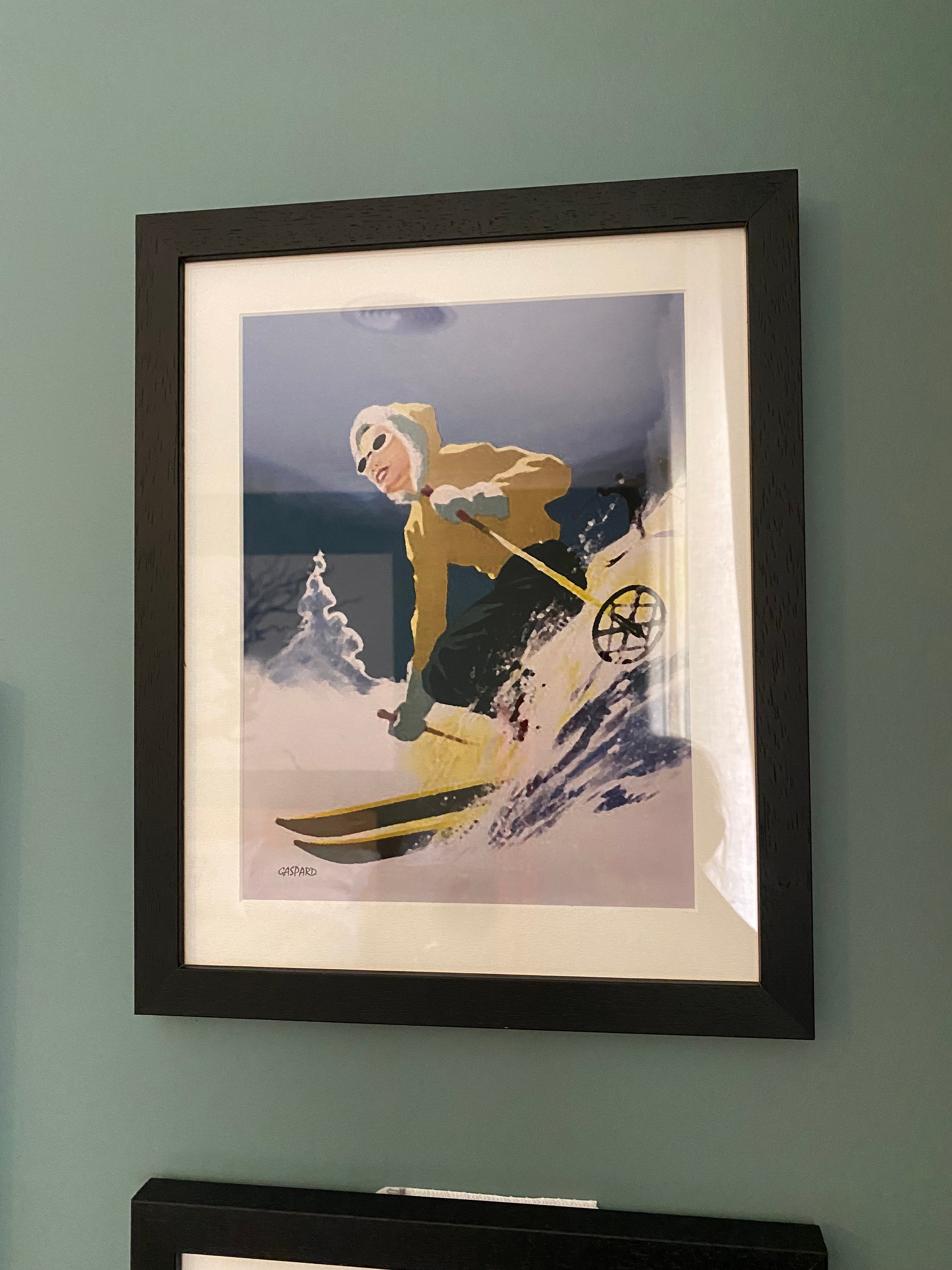 Black framed vintage photo of a woman in a yellow jacket with a white fur at the edge of the hood and blue pants skiing downhill, leaning to the right into the hill with a tree and the shadow of the mountains in the background below a blue sky, hanging on a green wall 