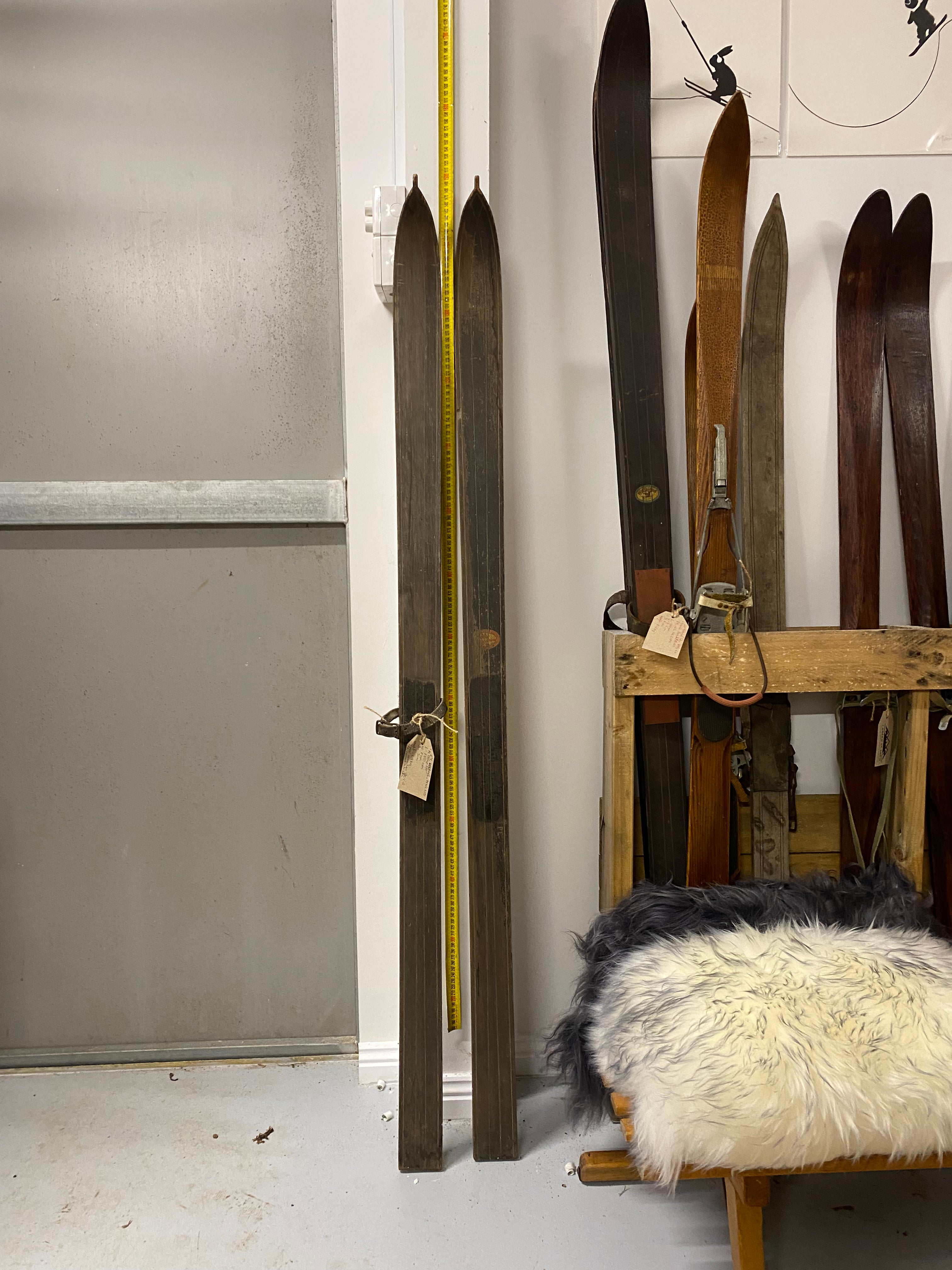 Full Height front view: Montgomery Ward vintage wooden skis. 1 skis with a leather strap. 1 ski sun faded. leaning against white painted wall with yellow measuring tape. More wooden skis stacked to the right hand side