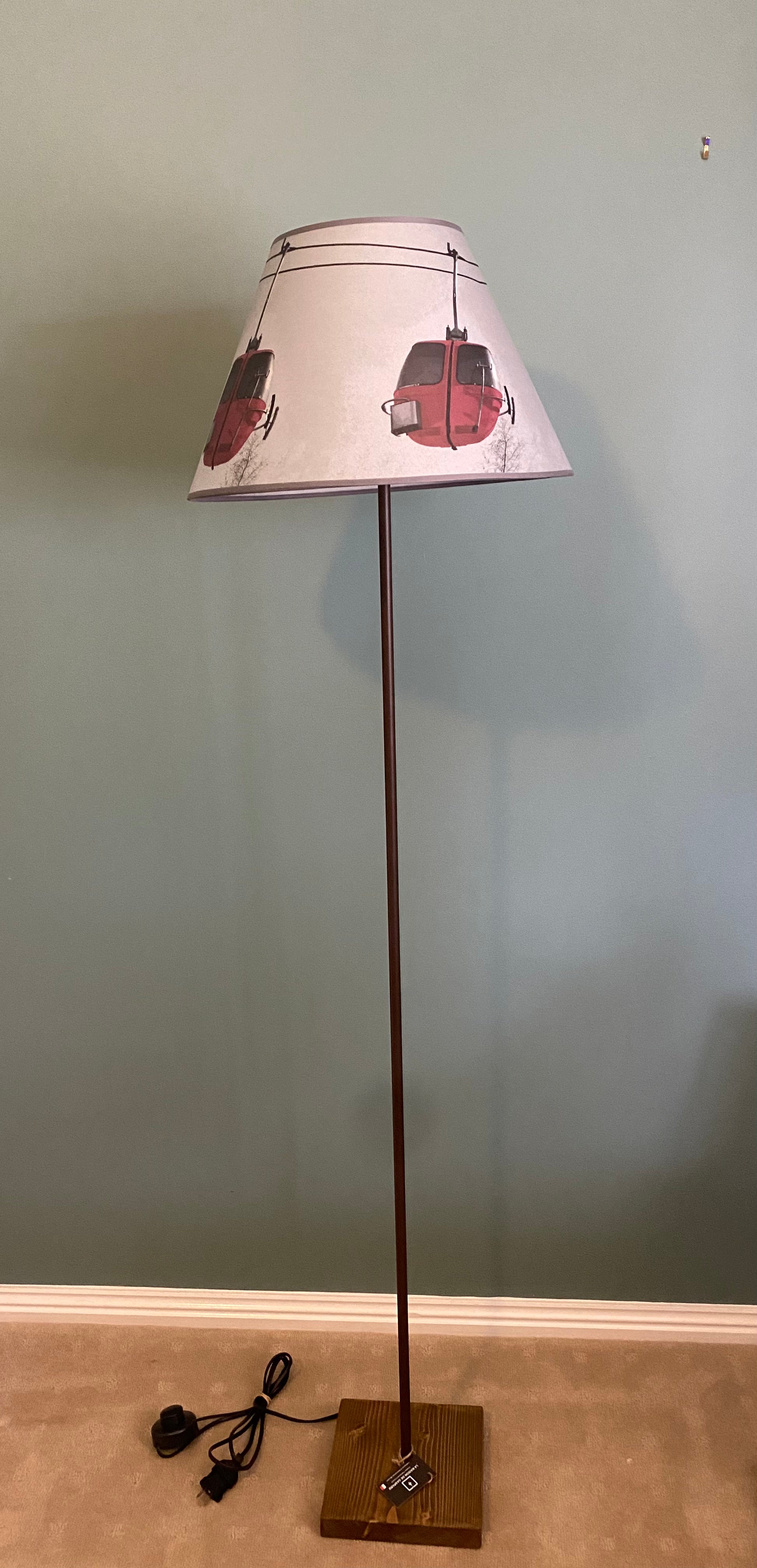 Photo of a floor lamp with a square solid wood base supporting a brown metal rod, topped by a vintage photo printed canvas lamp shade. The colour photo shows 3 red egg gondolas in the air with tree tops and clouds in the background against a grey sky. Lamp sits on beige carpet, in front of a green painted wall.