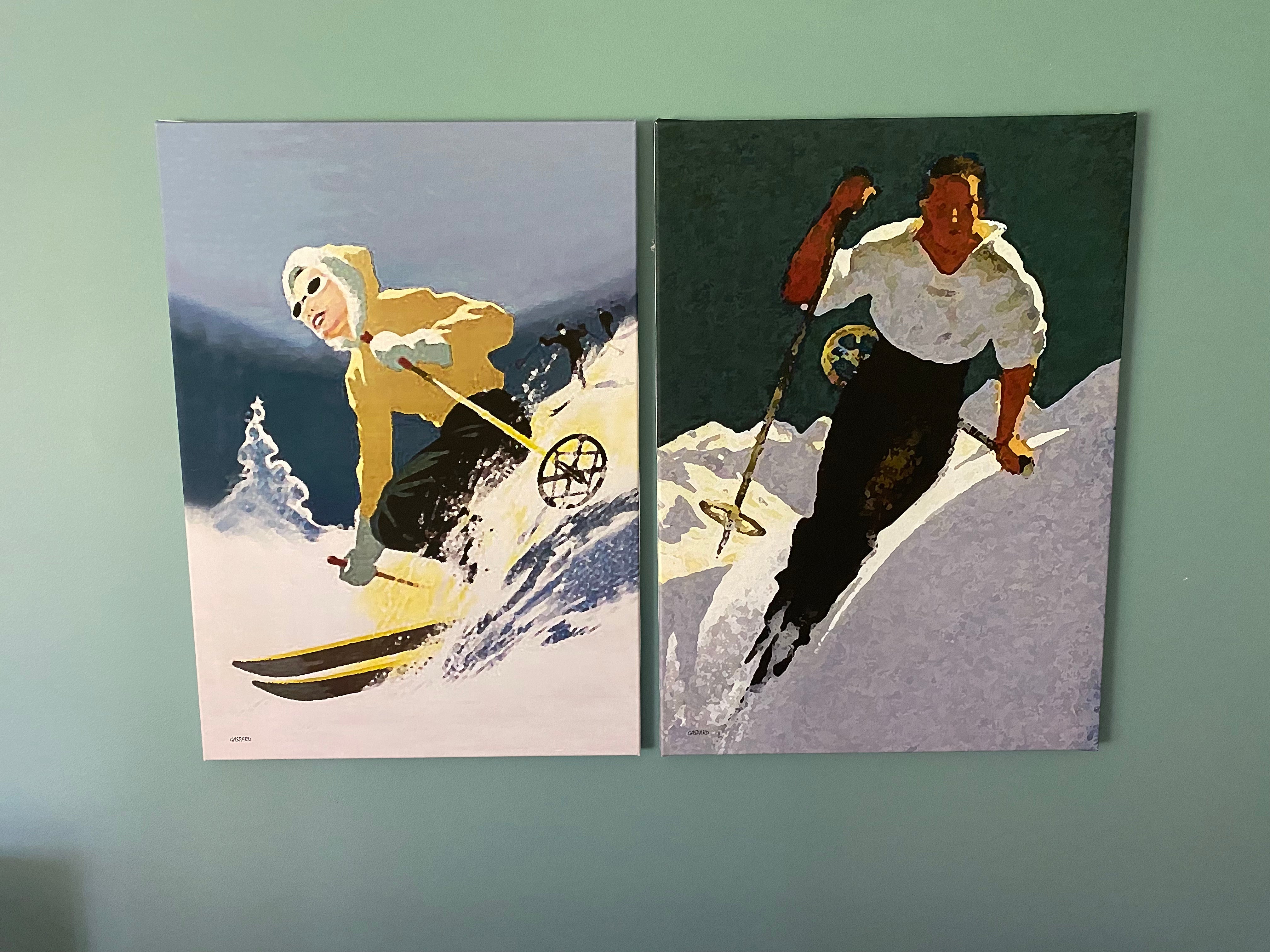 a man in a white shirt and blue pants skiing downhill, leaning left into the hill with mountains in the background below a dark green-blue tinged sky. Hanging next to a canvas depicts a woman in a yellow jacket with a white fur at the edge of the hood and blue pants skiing downhill, leaning to the right into the hill with a tree and the shadow of the mountains in the background below a blue sky. Hanging on a green wall.