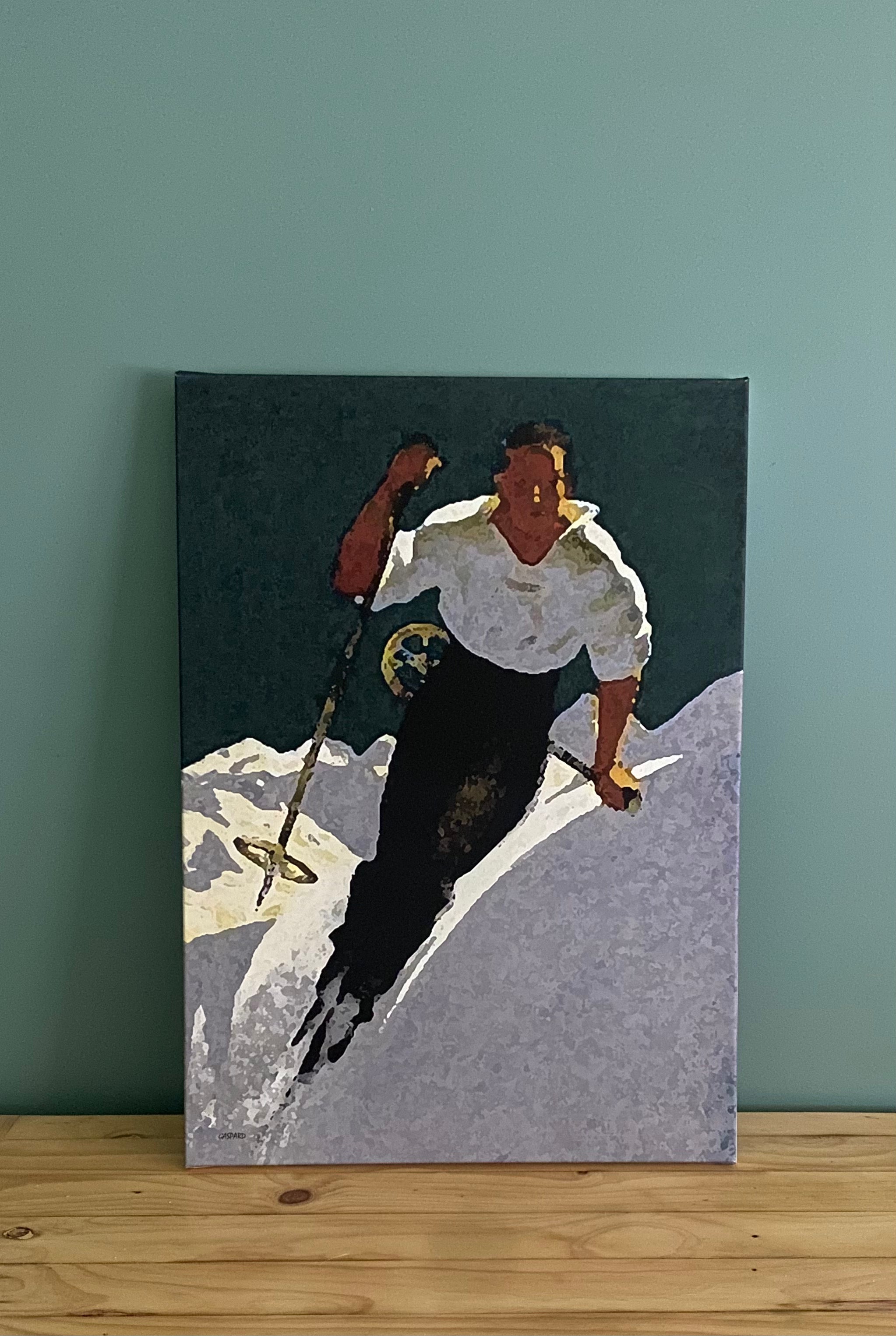 Canvas print of a man in a white shirt and blue pants skiing downhill, leaning left into the hill with mountains in the background below a dark green-blue tinged sky. Resting against a green wall on a timber table