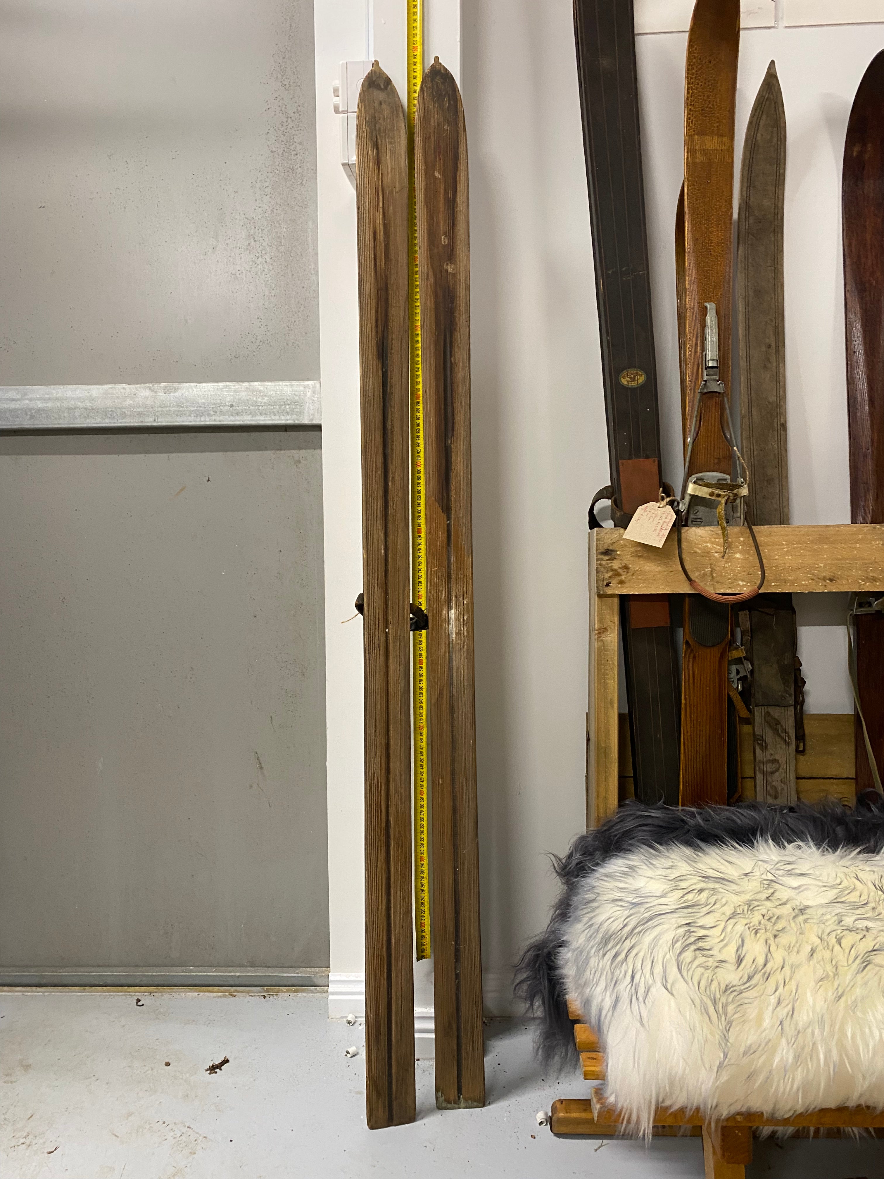 Full Height bases view: Montgomery Ward vintage wooden skis. Leaning against white painted wall with yellow measuring tape. Stacked to the right hand side are more wooden skis
