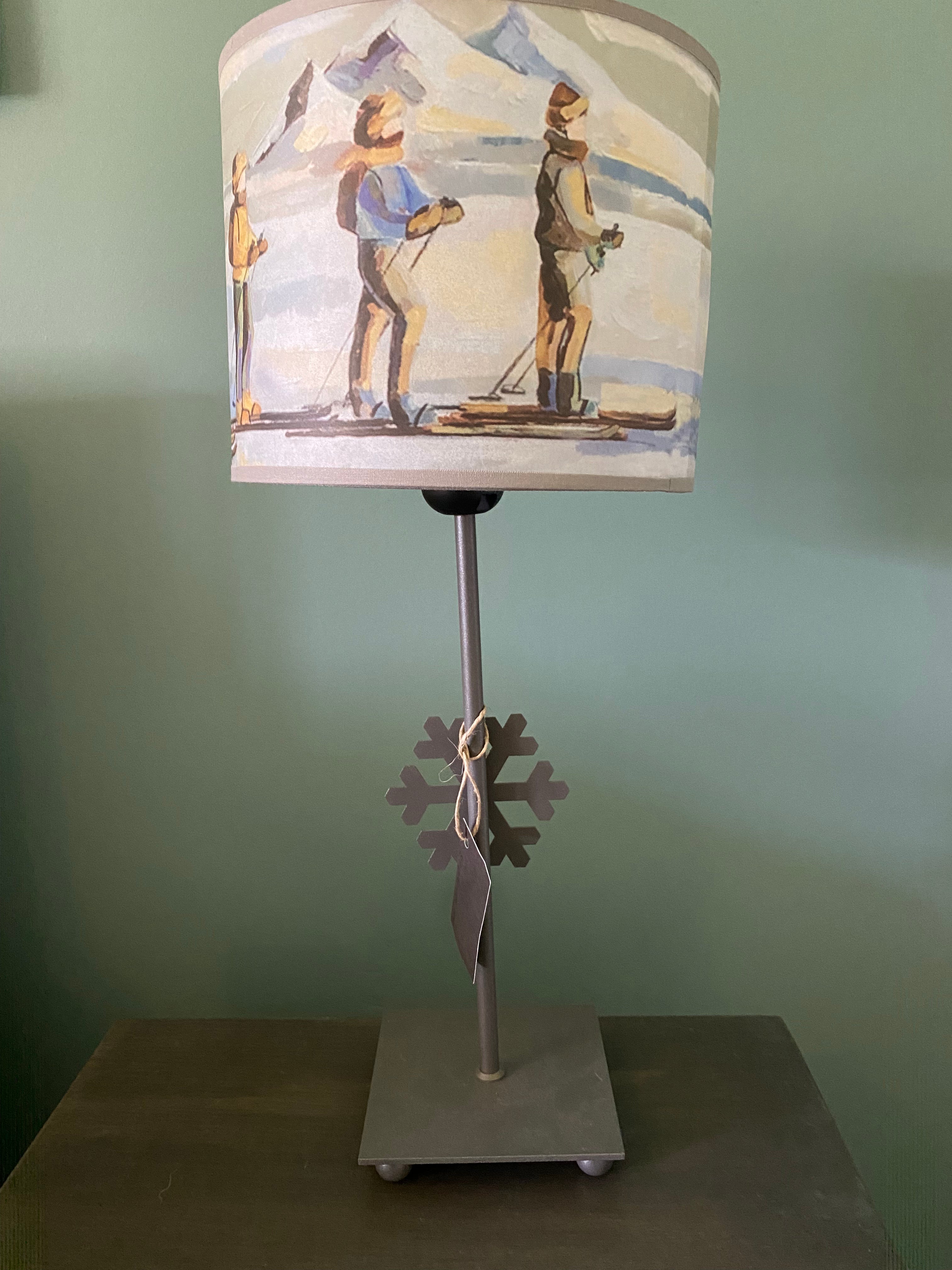 lamp with a square grey  solid metal base supporting a rod with ca snowflake mid way up the rod, topped by a colourful canvas lamp shade. The shade depicts people on skis skinning across the snow in a line, one in front of the other, with snow capped mountains in the background. Sitting on a  wooden table behind a green wall.