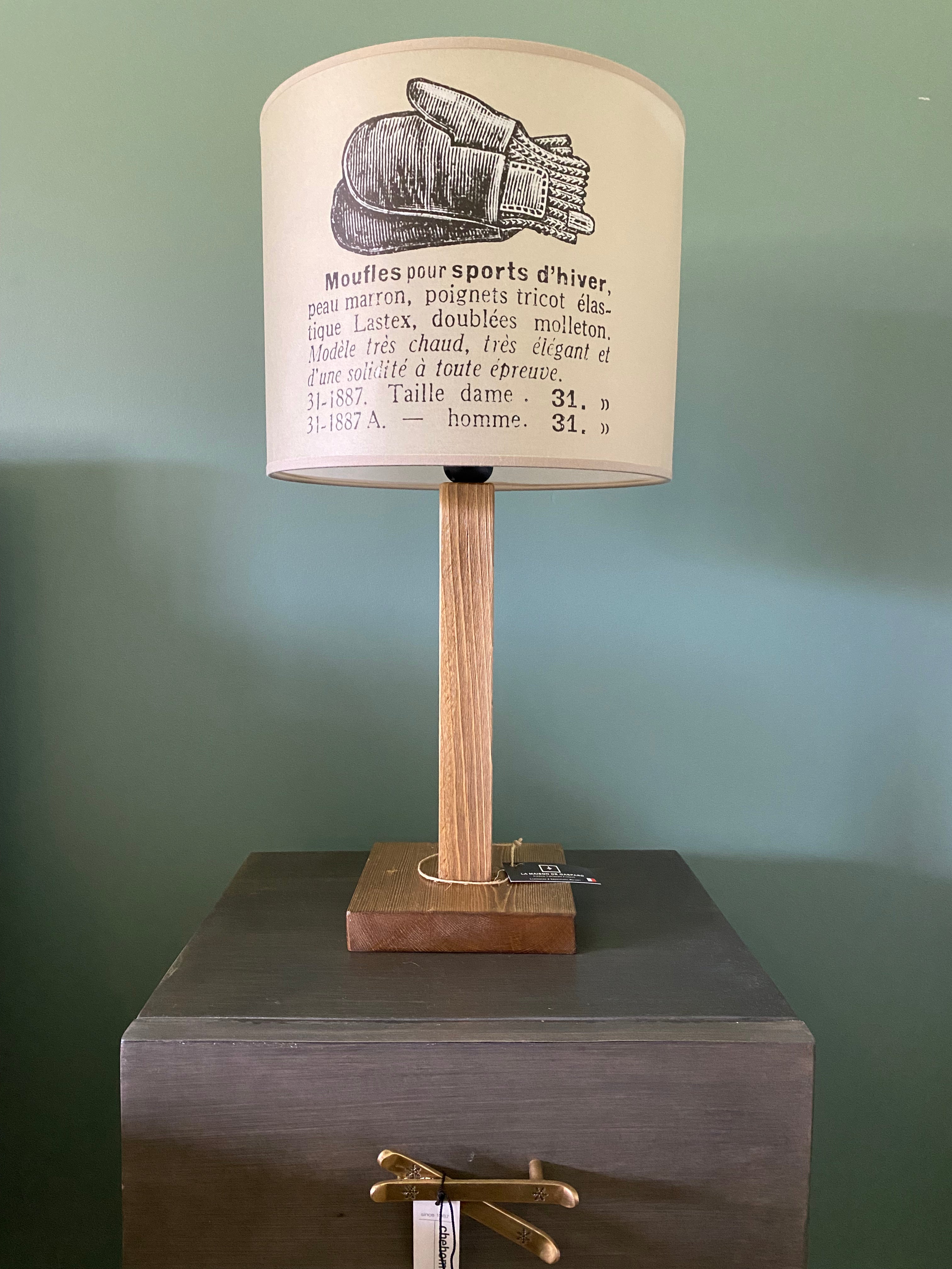 Lamp with a square solid wooden base and square stem holding up the canvas printed beige lamp shade. The beige shade shows a vintage black & white drawing of a pair of woollen mittens and description in french of the mittens. On a wooden table with brass ski handles against a green wall.