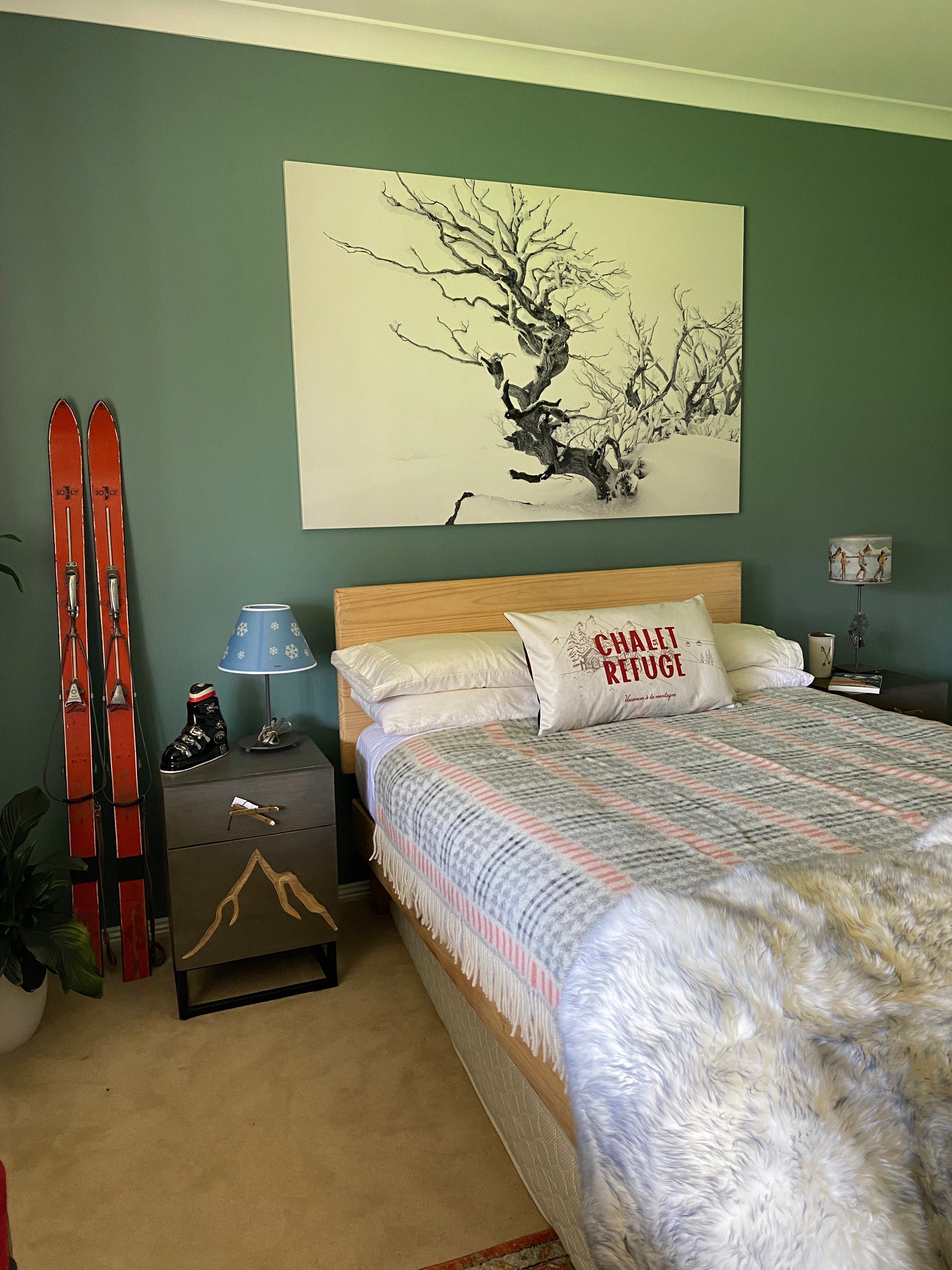 Wide angle view of a guest bedroom, with the Blue Snowflake lamp on the left hand bedside table. Room painted green, with vintage orange skis leaning against the wall. Grey, red & white blanket on the double bed and one throw cushion and a grey tipped white long-hair sheepskin rug.