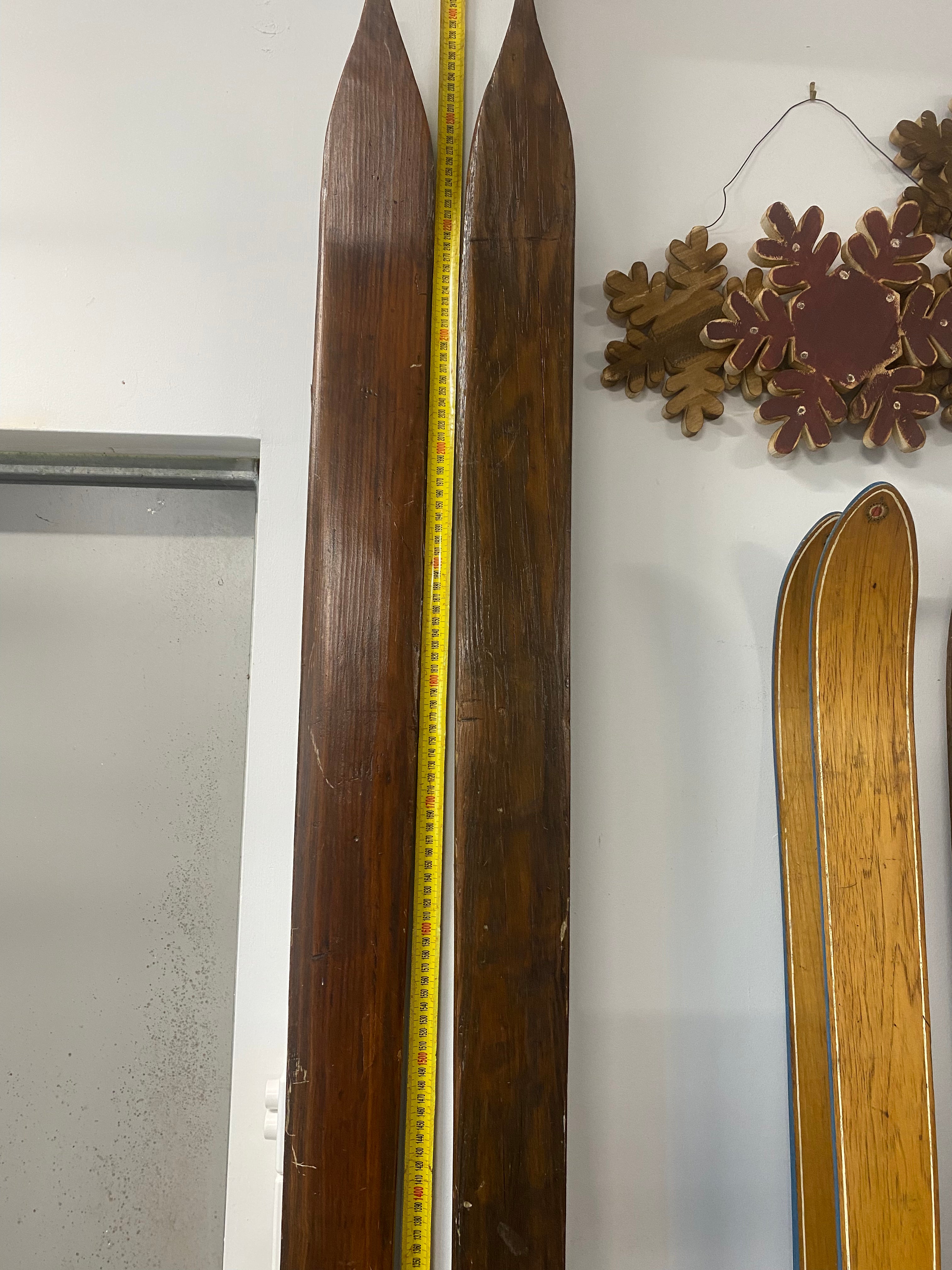 Vintage Wooden Skis: top third of both skis on white wall and resting against a yellow measuring tape.