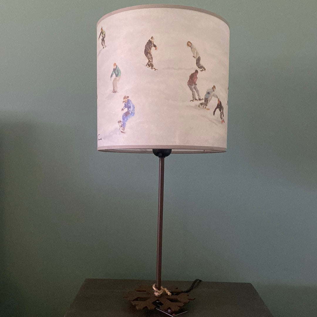 lamp with a round brown metal base in the shape of a snowflake, supporting a metal rod, topped by colour printed canvas lamp shade depicting a slope full of snowboarders riding downhill, in front a green wall & on top of a wooden table.