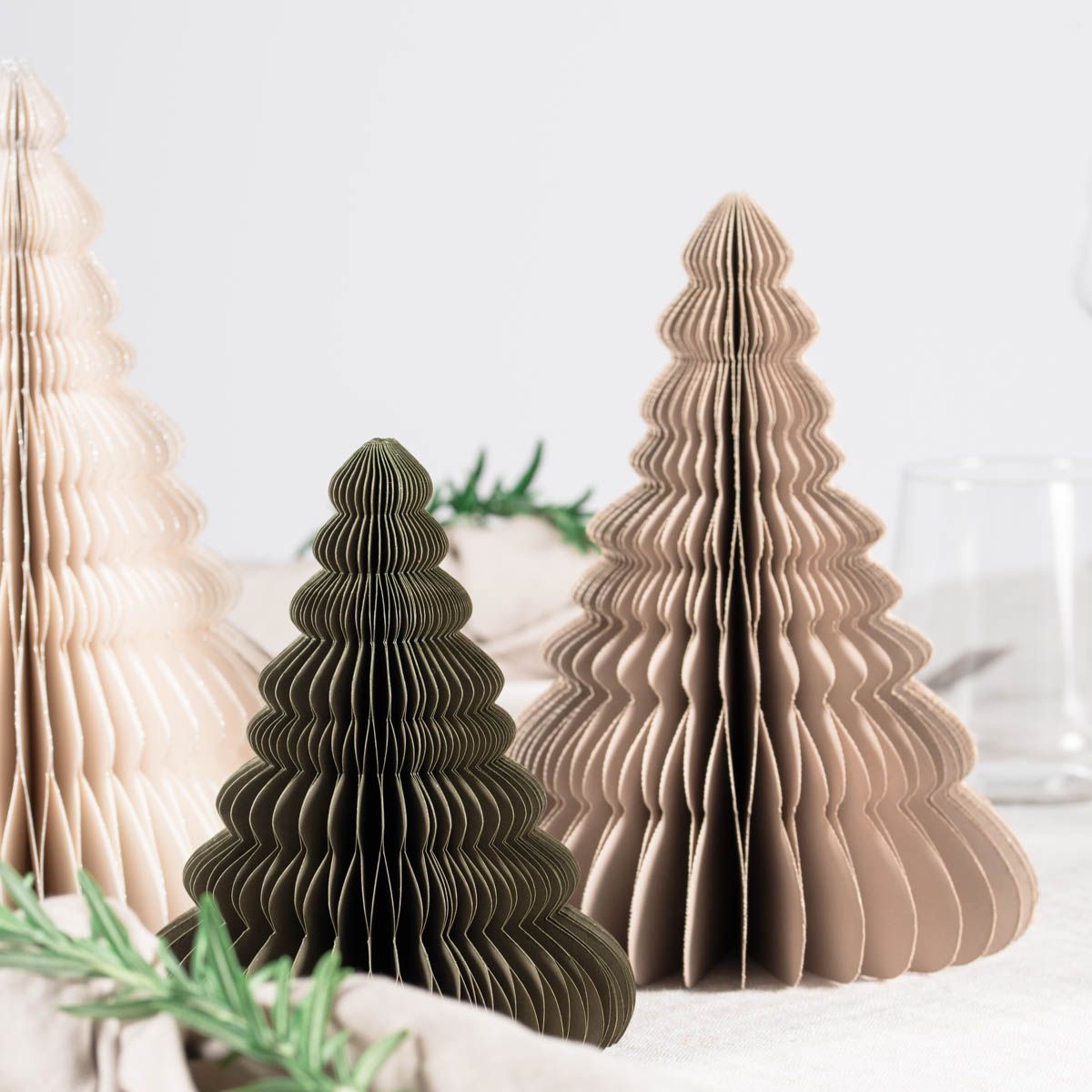 1 Off-White, 1 Olive Green  and 1 Linen coloured Paper Standing Christmas Trees, in front of a white painted wall and resting on a table laid with white linen with glassware and rosemary sprigs