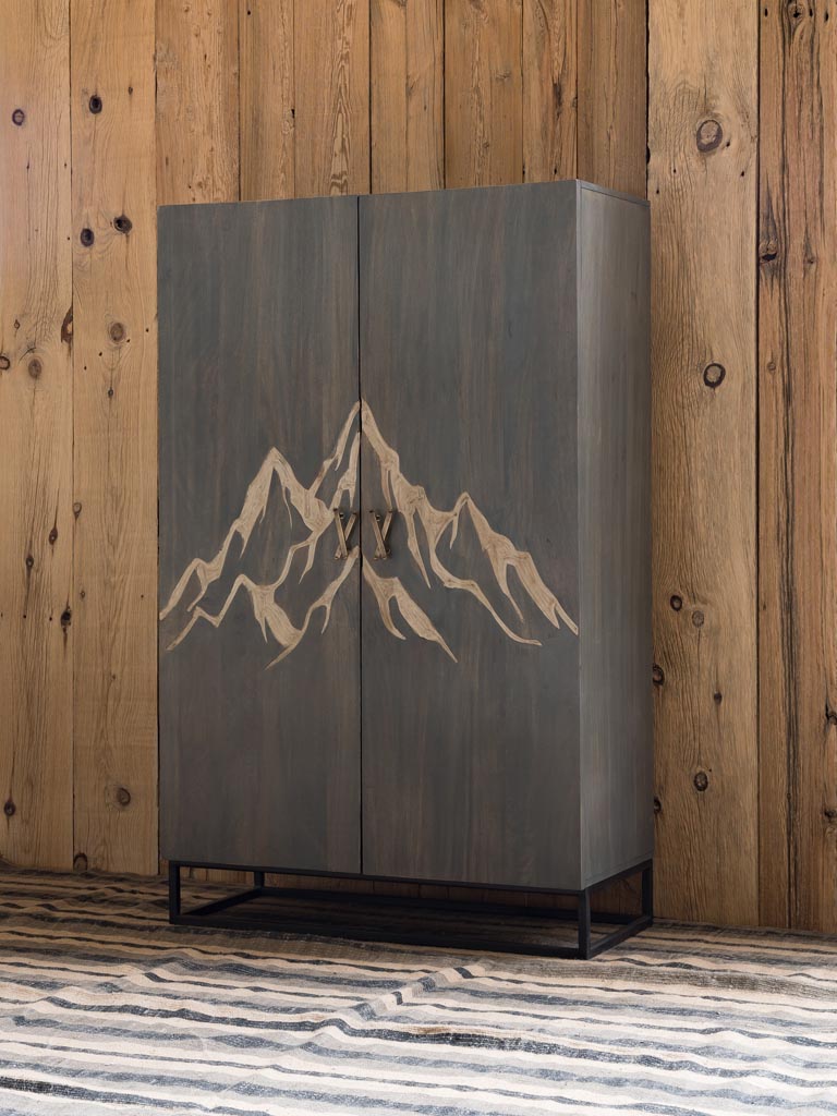 Side view: Wooden Cabinet engraved with a mountain & brass ski shaped handles with square metal base & legs on a wood panel background with rug underneath