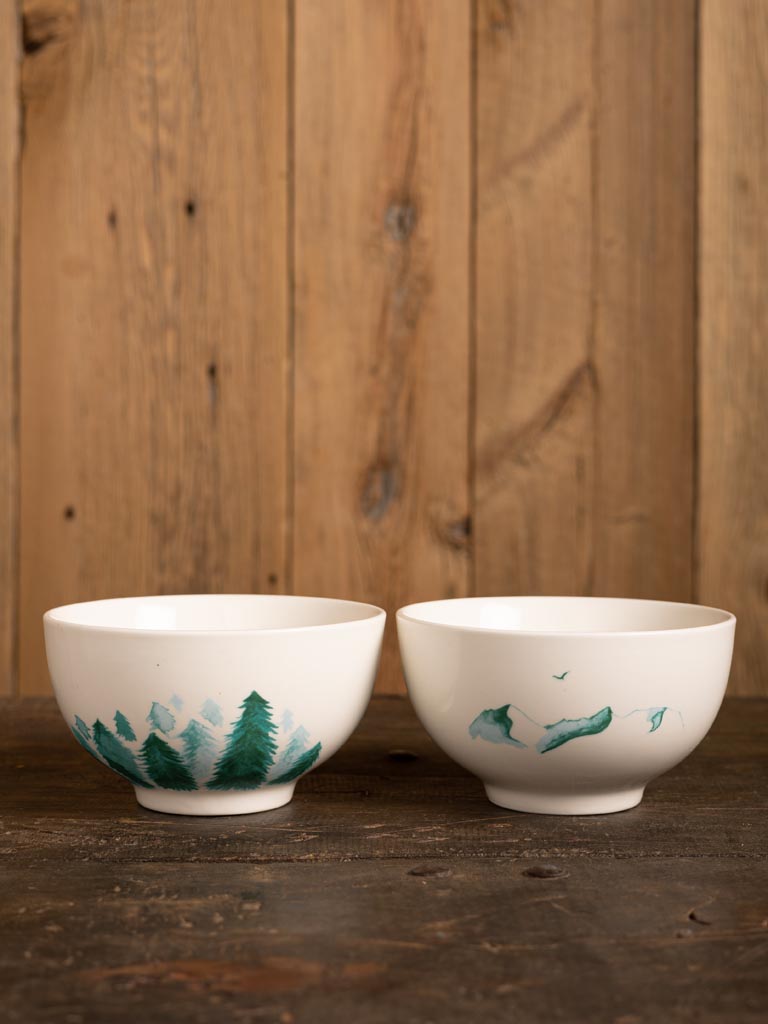 Pair of white porcelain bowls painted with trees and mountains in green, against a wood panelling wall and floor