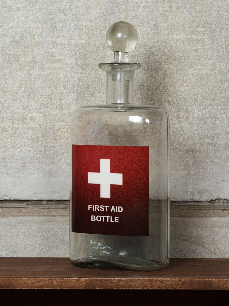 Glass Decanter with the red and white First Aid symbol labels on its front & with glass stopper, on a wood table and against a  light grey wall