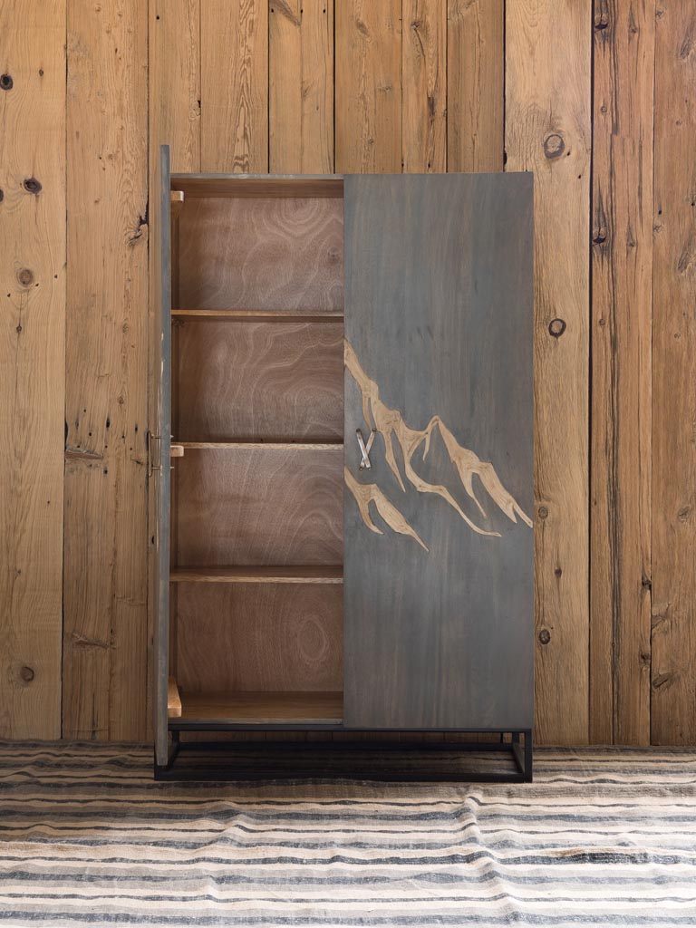 1 Cabinet door open showing internal wood shelves: Wooden Cabinet engraved with a mountain & brass ski shaped handles with square metal base & legs on a wood panel background with rug underneath