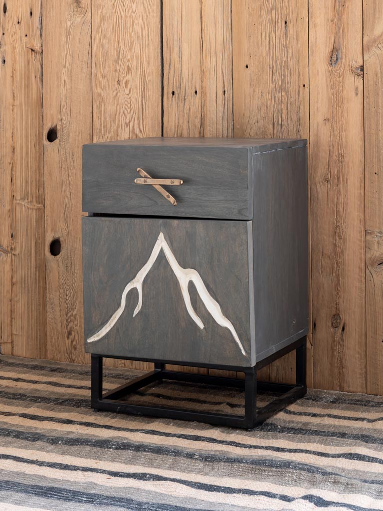 Side view of a Bedside table engraved with a mountain & ski handle with a wood panelled wall in the background and a rug on the floor