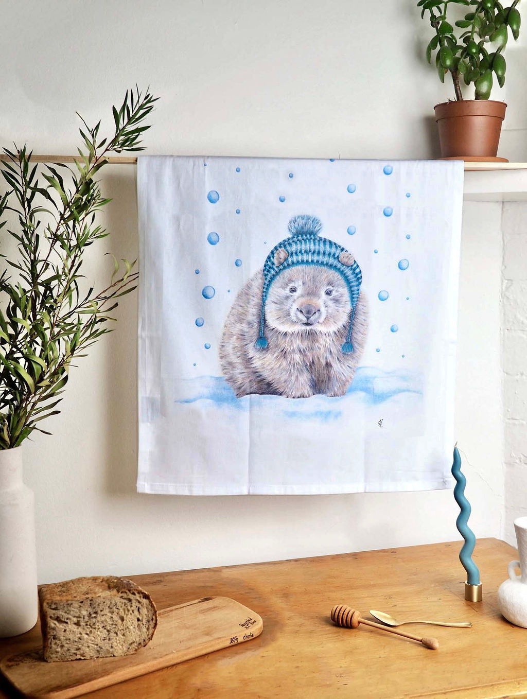 Image shows a tea towel illustrated with a wombat wearing a blue tasseled beanie, standing in blue tinged snow while the snow falls. The tea towel is hanging on a wall using a pale wooden hanging rail above a wooden kitchen bench and against a white kitchen backdrop.
