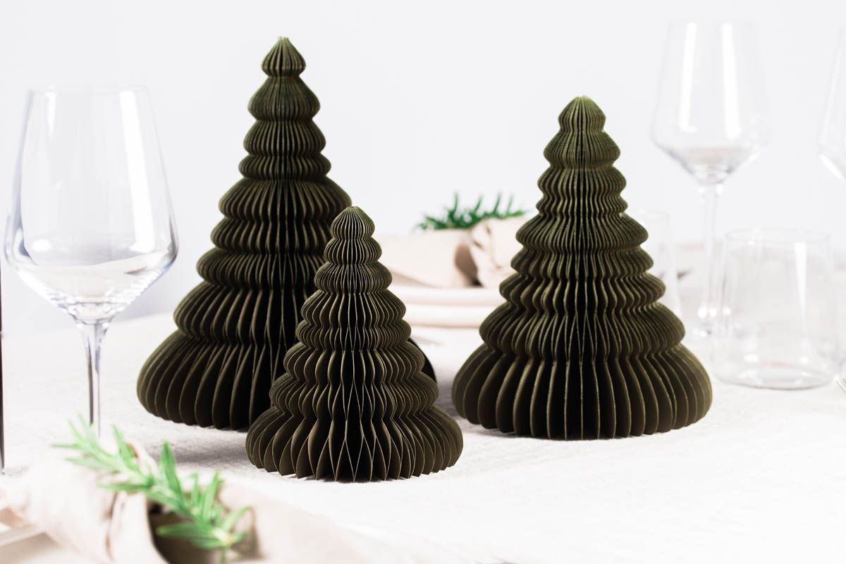 Selection of 3 Olive Green Paper Standing Christmas Trees, in front of a white painted wall and resting on a table laid with white linen with glassware and rosemary sprigs